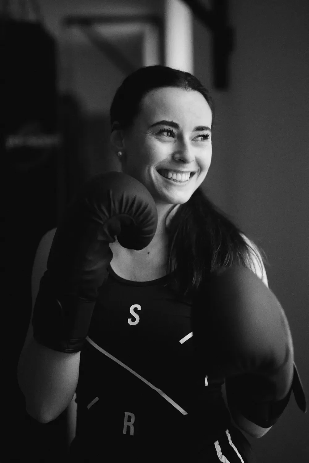 Schwarze und weiße Aufnahme einer lachenden jungen Frau, die Handschuhe trägt und bereit für Boxtraining ist.