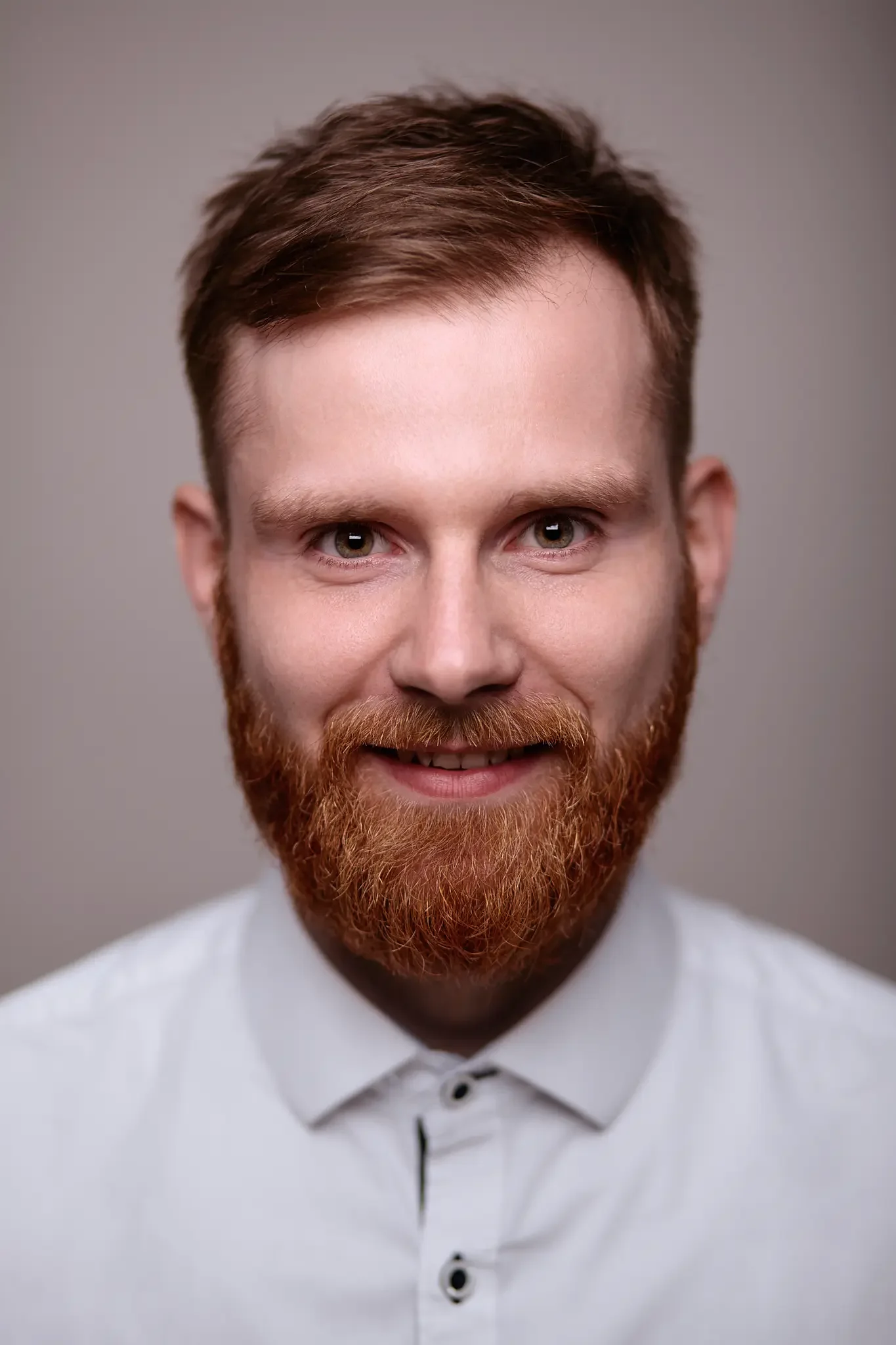 Porträt eines Mannes mit rotem Bart, kurzen braunen Haaren und weißem Hemd vor grauem Hintergrund.