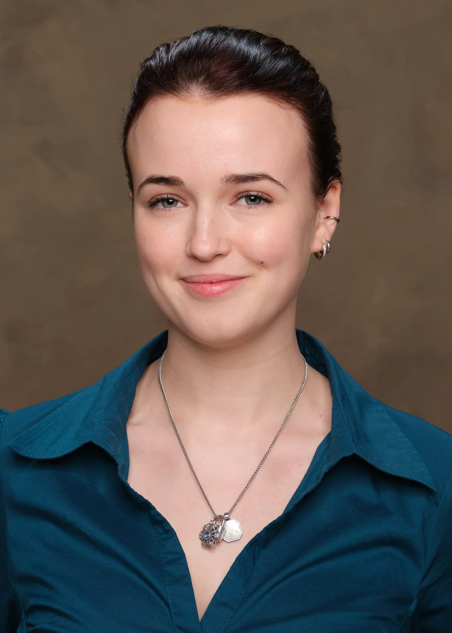 Porträt einer Frau mit dunklen Haaren, blauer Bluse und Schmuck, vor braunem Hintergrund.