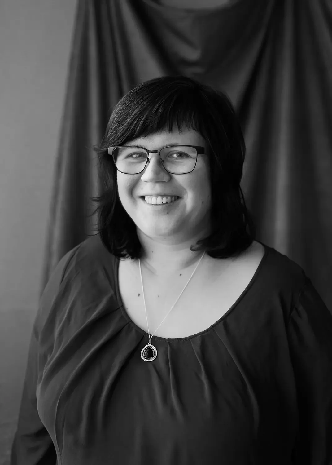 Schwarz-Weiß-Foto einer lächelnden Frau mit Brille, dunklem Haarschnitt, schwarzen Oberteil und silberner Halskette vor dunklem Vorhang.