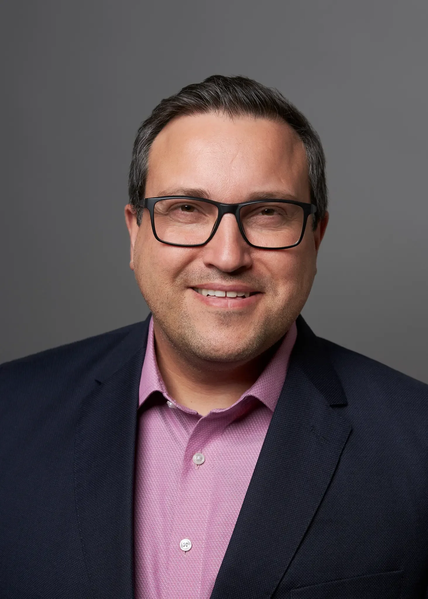 Porträt eines lächelnden Mannes mit dunklen Haaren, Brille, Hemd und Jacke vor grauem Hintergrund.