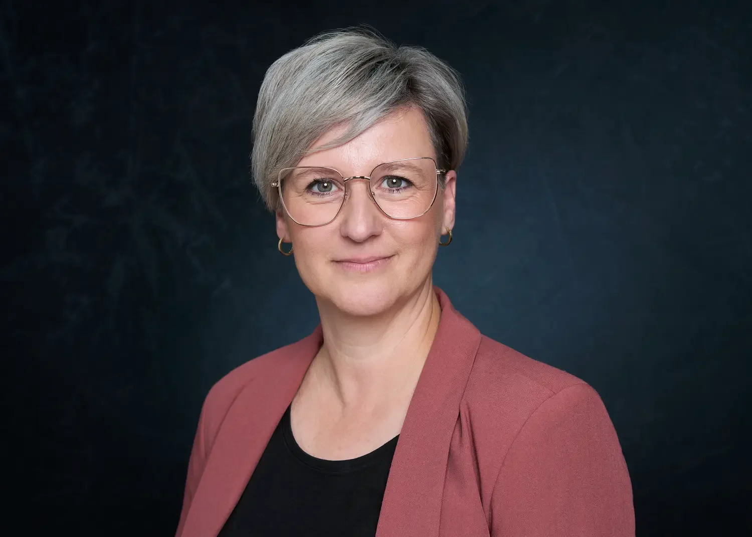 Porträt einer älteren Frau mit kurzen grauen Haaren, Brille, in schwarzem Top und rosa Jacke, vor dunklem Hintergrund.
