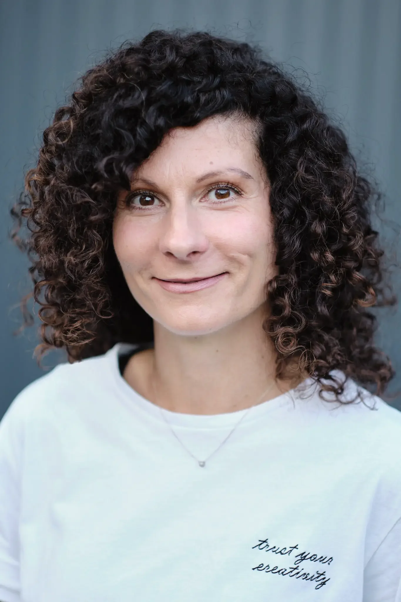 Nahaufnahme einer lächelnden Frau mit lockigem dunklem Haar vor einem grauen Hintergrund, trägt ein weißes T-Shirt mit der Aufschrift "trust your creativity" und eine zarte Halskette.