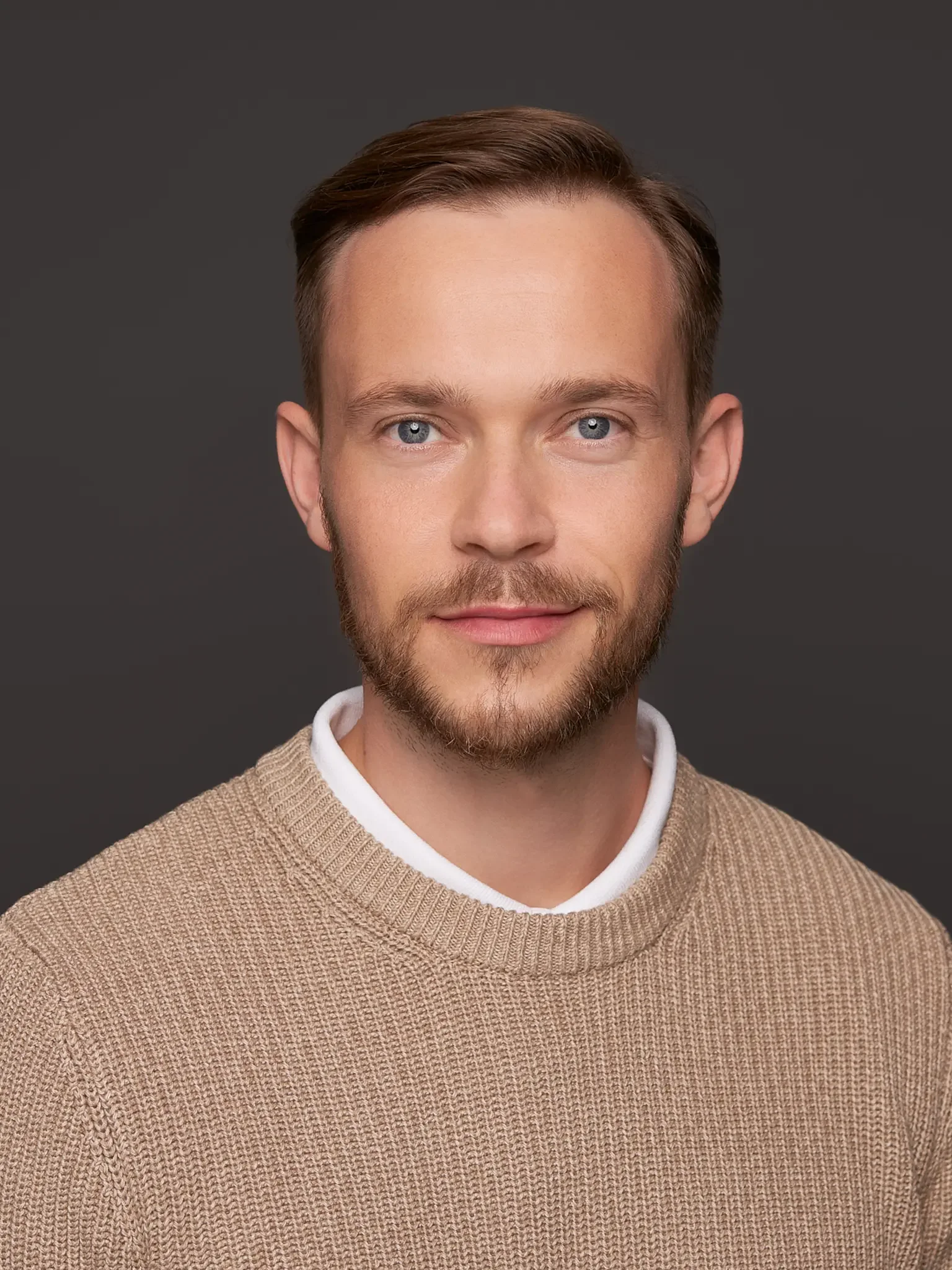 Porträt eines jungen Mannes mit braunem Haar und Bart, trägt einen hellbraunen Pullover vor einem grauen Hintergrund.