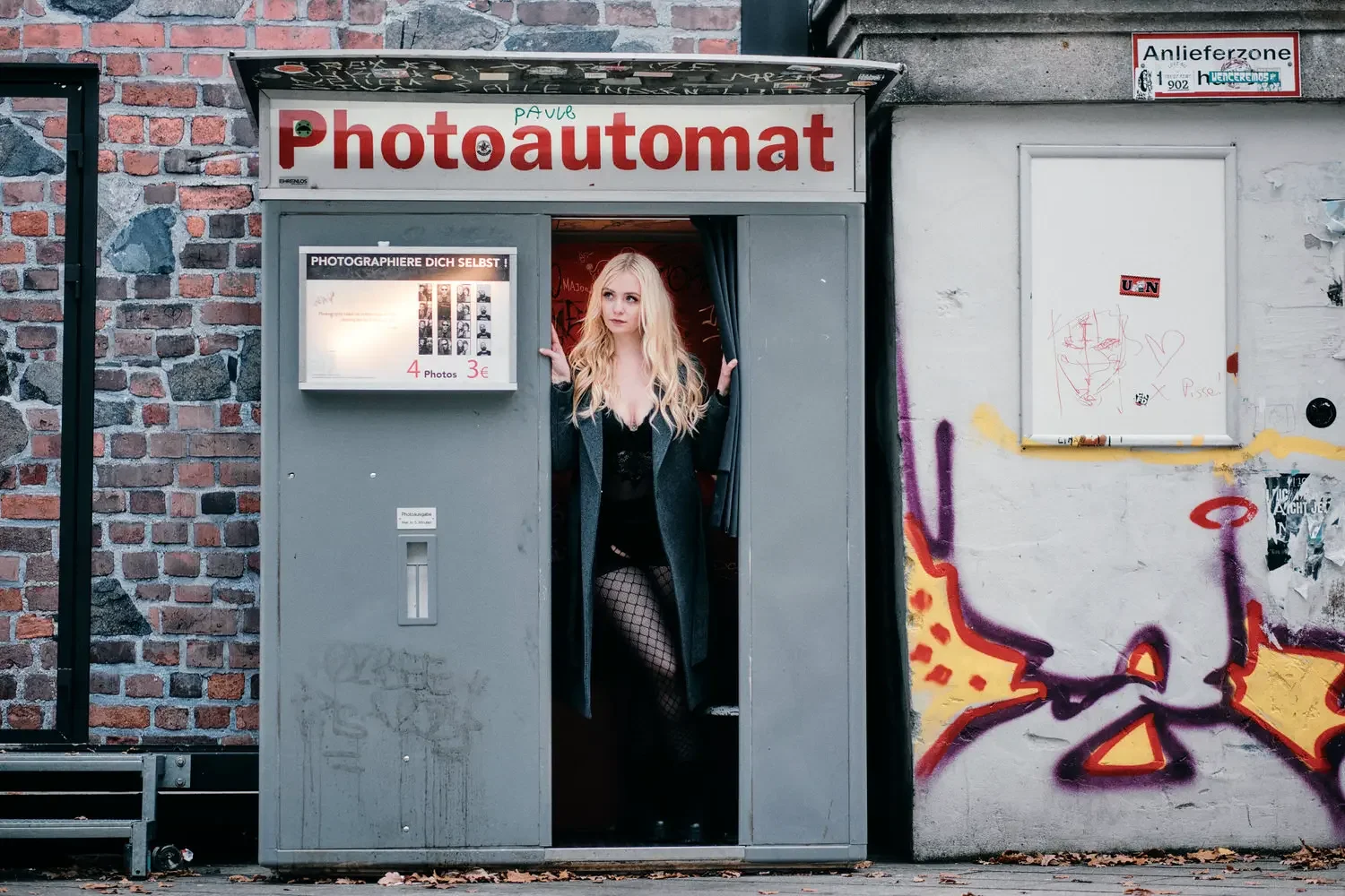 Junge Frau in schwarzem Kleid und Netzstrümpfen steht in einer Fotokabine mit roten Vorhängen, die an einer Stadtmauer mit Graffiti befindet.