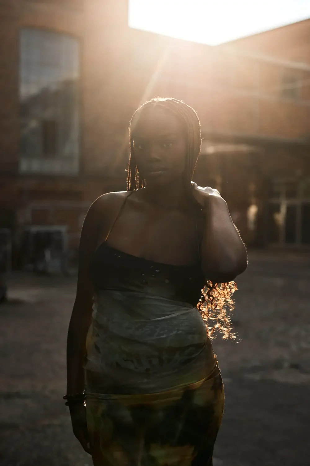 Frau mit lockigem Haar in einem schwarzen Oberteil, die in der Sonne steht, vor einem urbanen Hintergrund mit Mehrfamilienhäusern.