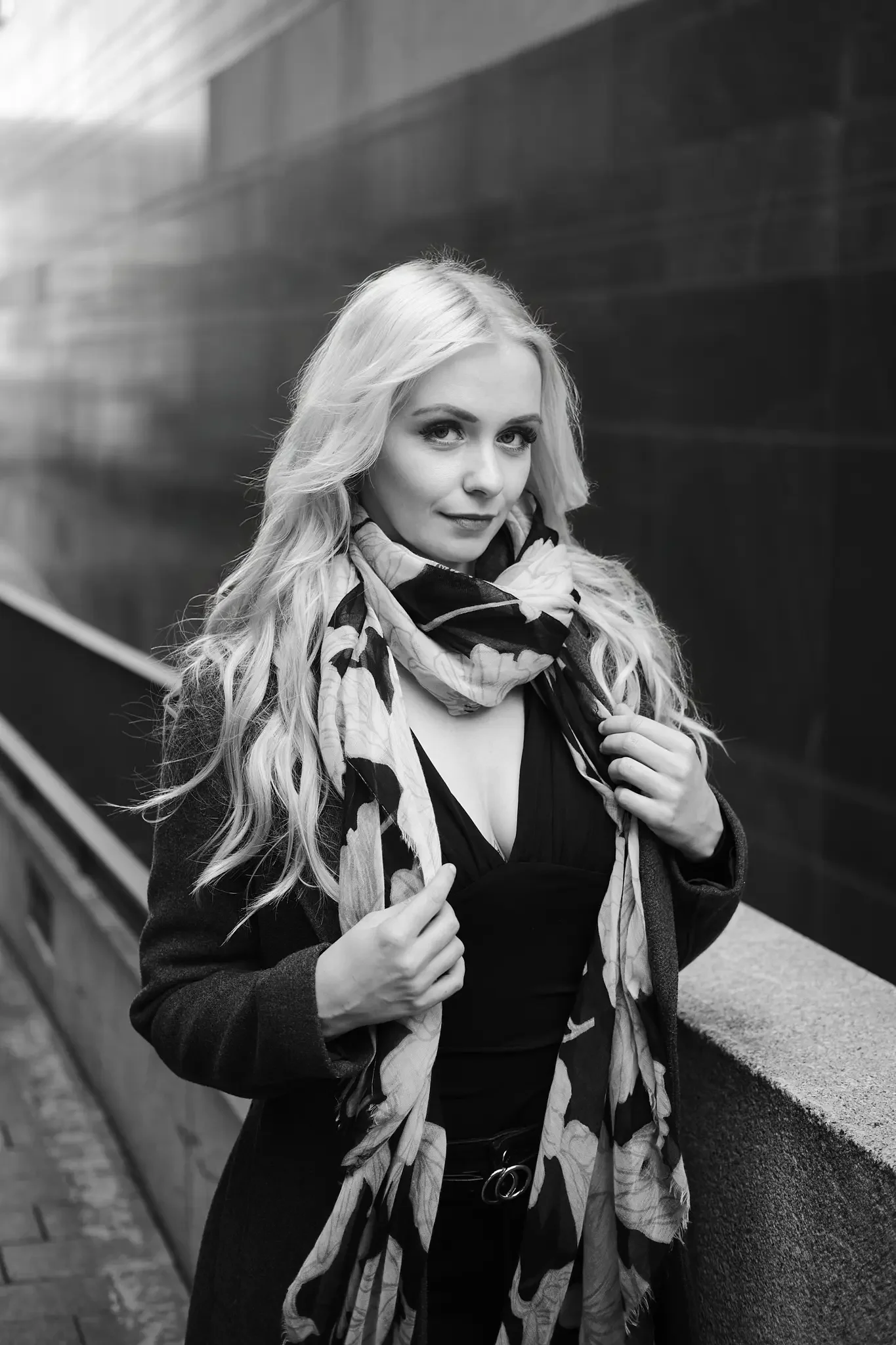 Porträt einer jungen blonden Frau mit lockigem Haar, die an einer Betonmauer steht, trägt eine dunkle Jacke, ein mit Blumen bedruckter Schal und einen Gürtel mit Doppelringe, in einer Stadtumgebung.