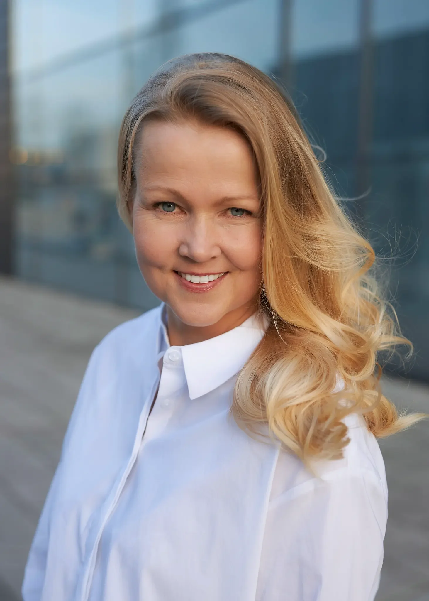 Nahaufnahme einer lachenden Frau mit lockigem, blondem Haar, die weiße Bluse trägt, vor einer modernen Gebäudefassade im Freien.