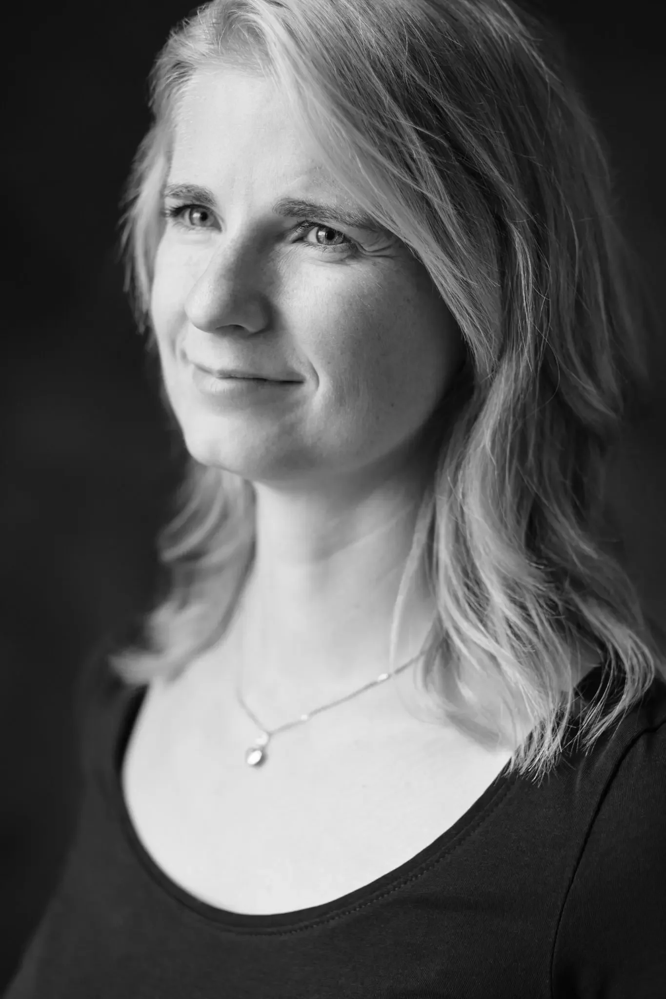 Schwarz-weiß Porträt einer lächelnden Frau mit Schulterlänge haaren und einer Halskette, im Vordergrund. 