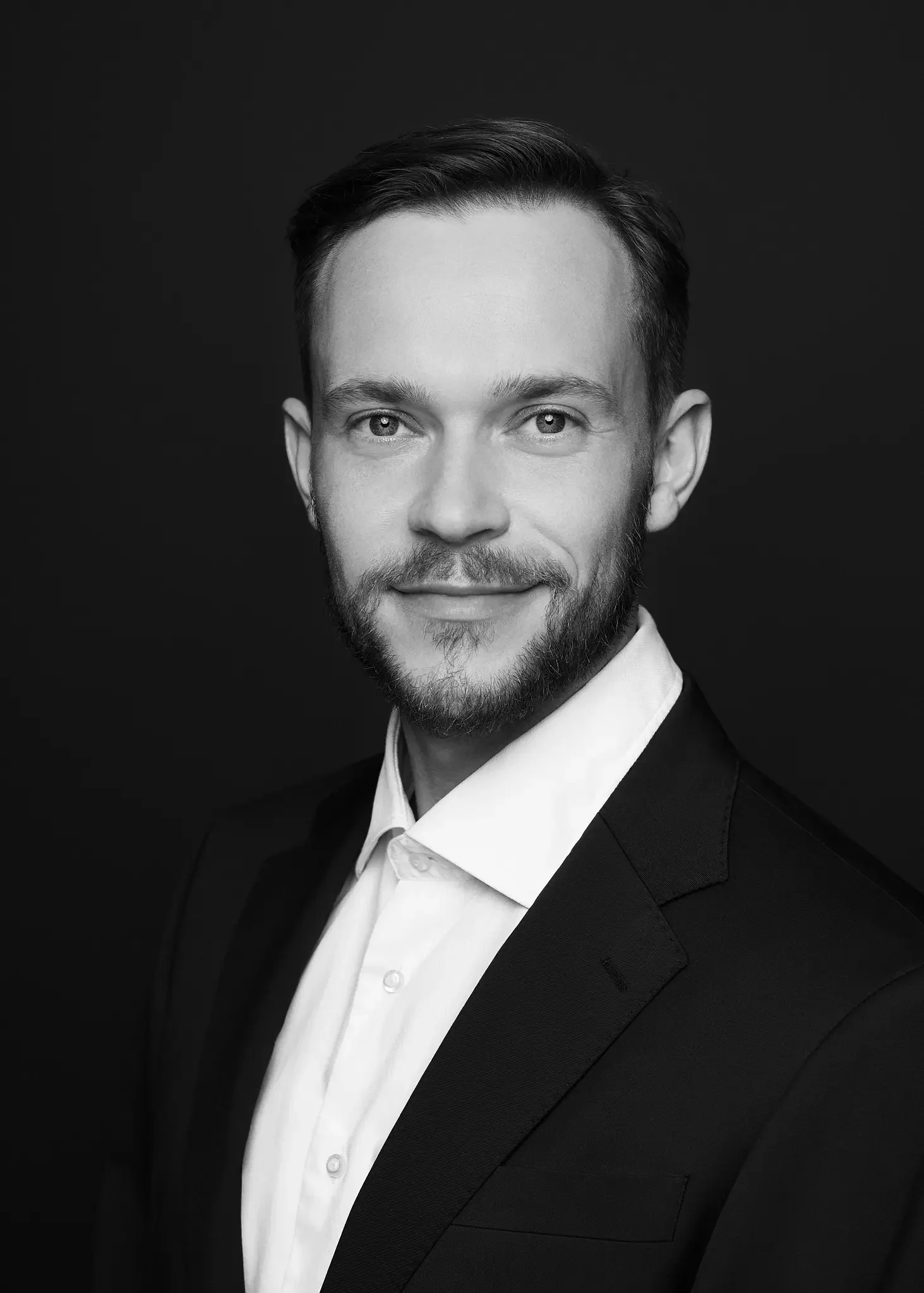 Schwarz-weiß-Porträt eines jungen Mannes in formeller Kleidung, lächelnd, vor dunklem Hintergrund.