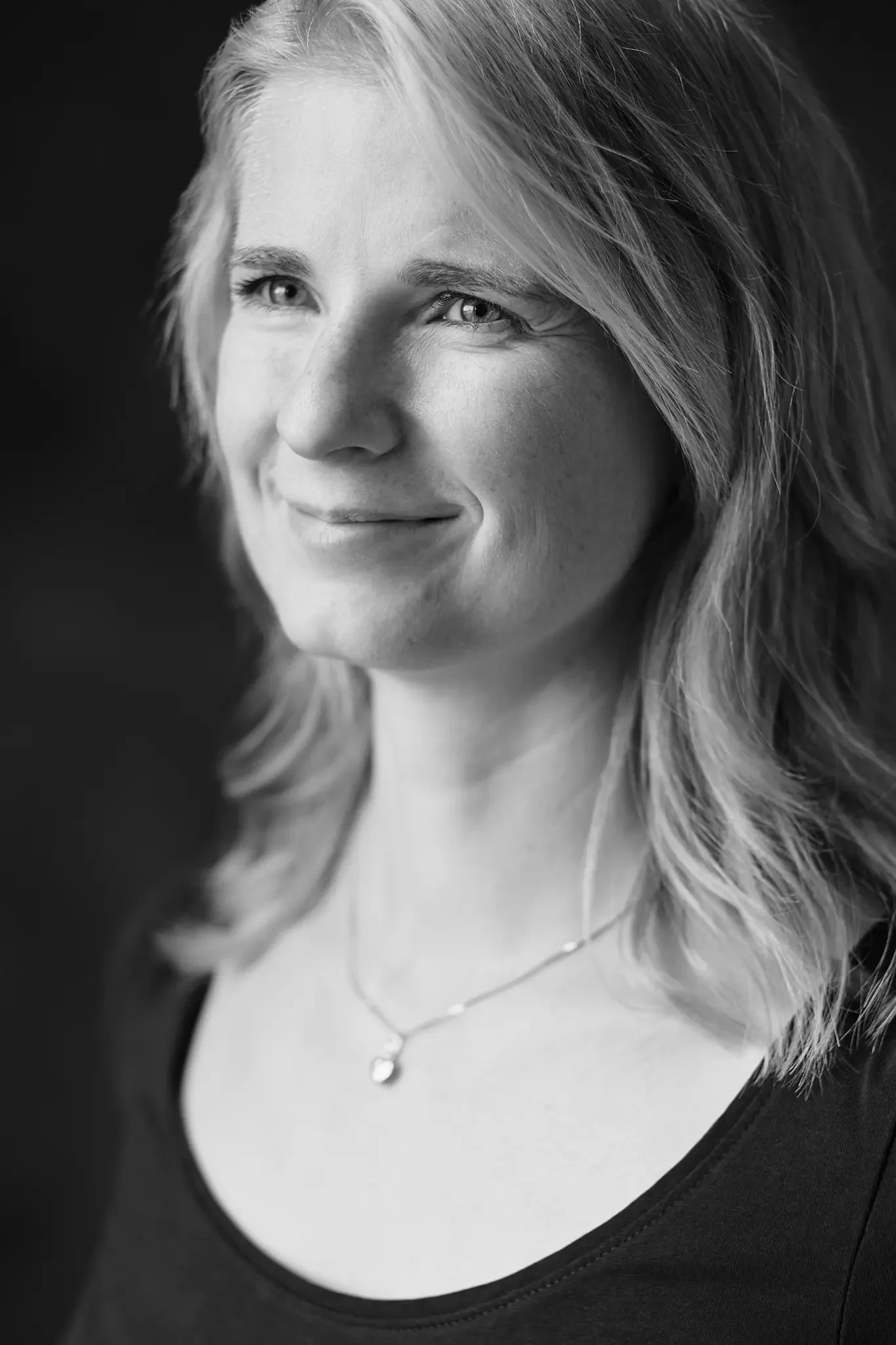 Schwarz-Weiß-Porträt einer lachenden Frau mit lockigem, schulterlangem Haar und Halskette, lächelnd, vor dunklem Hintergrund.