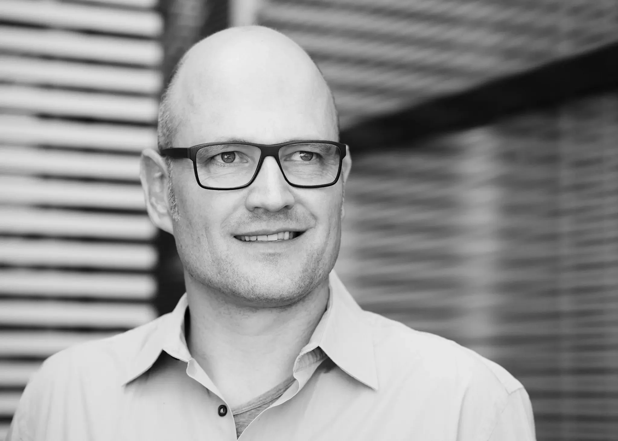 Schwarz-Weiß-Foto eines lächelnden Mannes mit Brille, hemden und Glatze vor einem modernen, vertikal gestalteten Hintergrund.