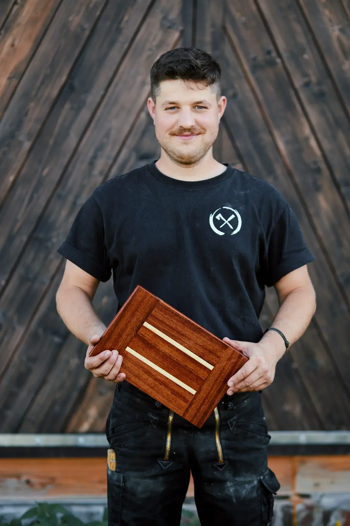 Mann hält handgefertigtes Holzbrett vor einer dunklen Holzwand.