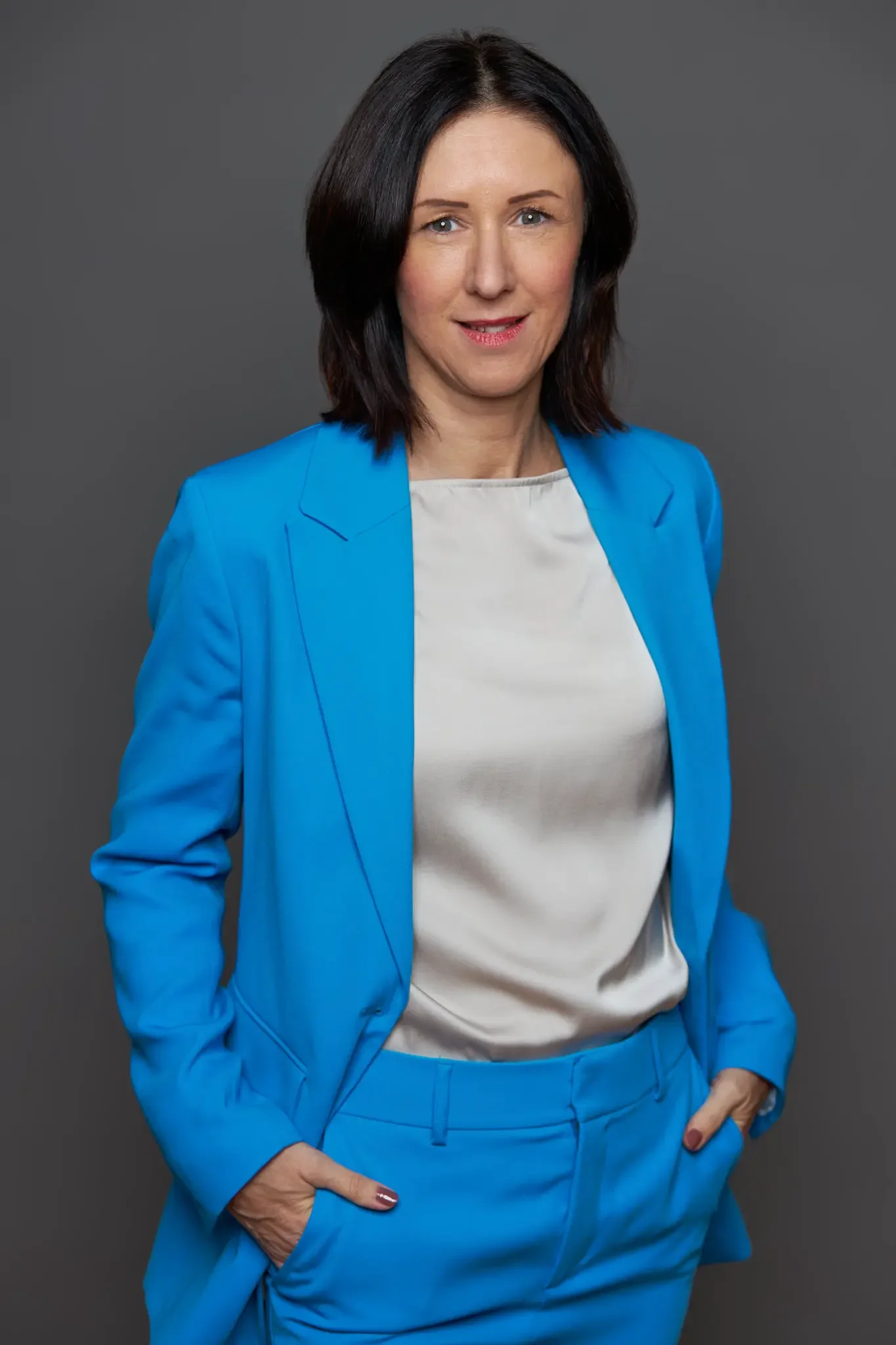 Eine Frau mit dunklen, schulterlangen Haaren trägt einen blauen Anzug und ein cremefarbenes Oberteil, steht vor grauem Hintergrund und lächelt leicht.