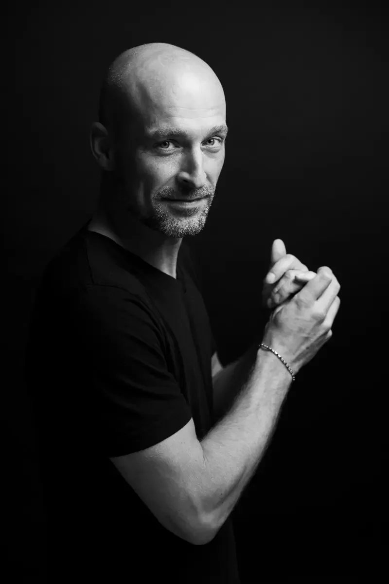 Schwarz-weiß-Porträt eines blonden Mannes mit Glatze, der in die Kamera schaut, die Hände zusammengekniffen und ein Lächeln zeigt.