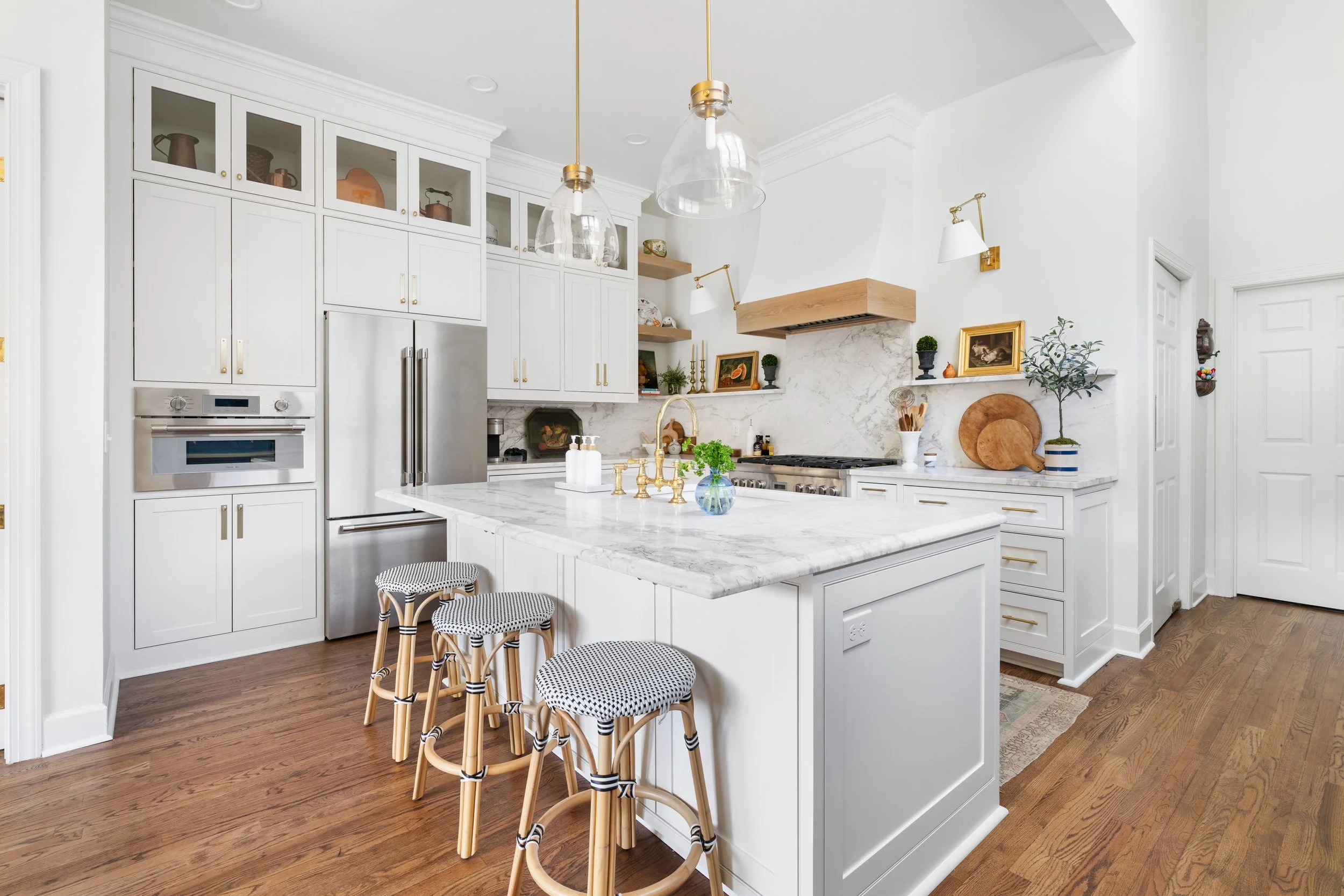 Bright white kitchen with marble countertops, wooden stools with woven seats, stainless steel refrigerator, and decorative shelves with art and plants.