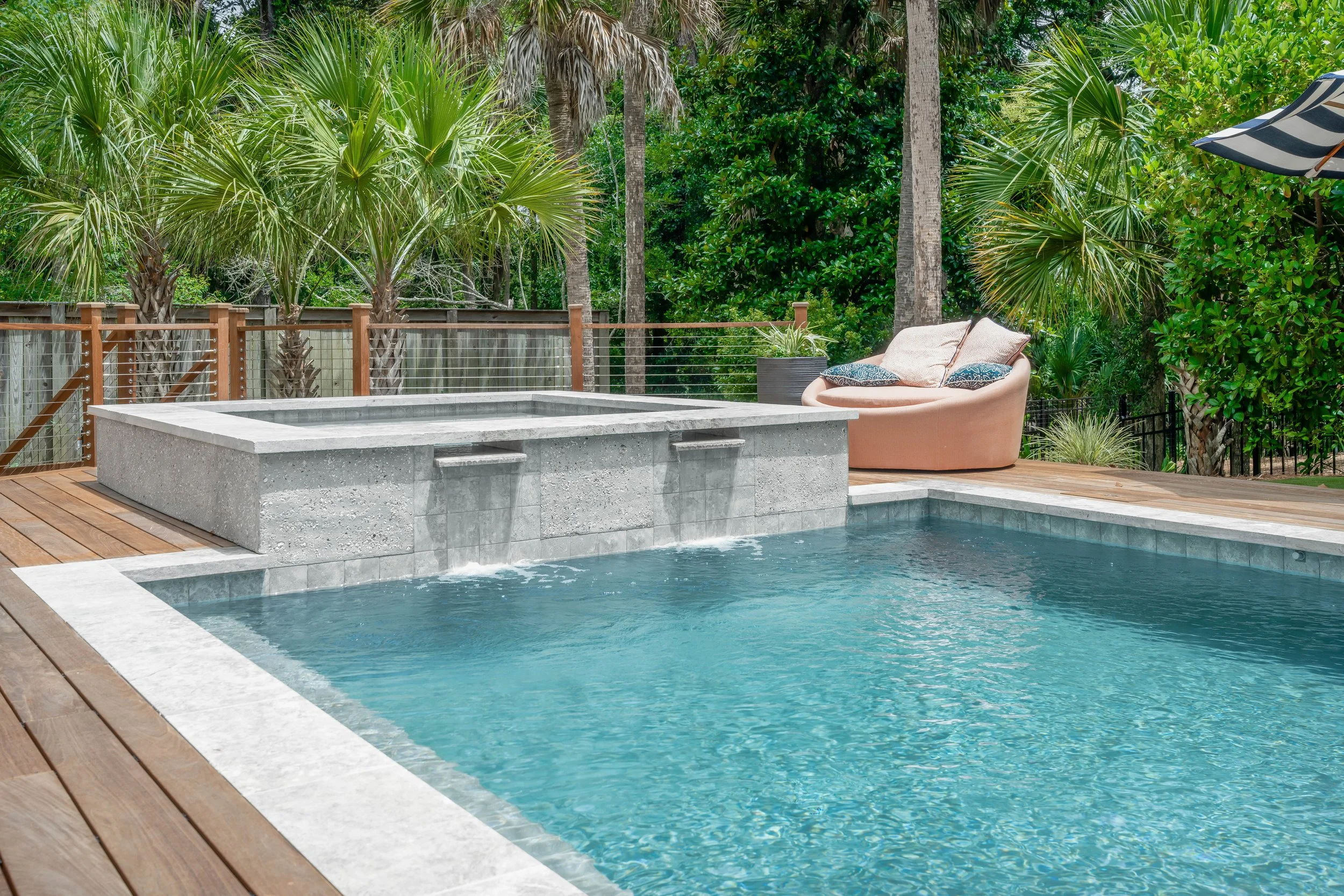 A backyard swimming pool with a stone hot tub attached, surrounded by wooden decking and lush green tropical trees and plants, with a pink outdoor sofa and throw pillows in the background.