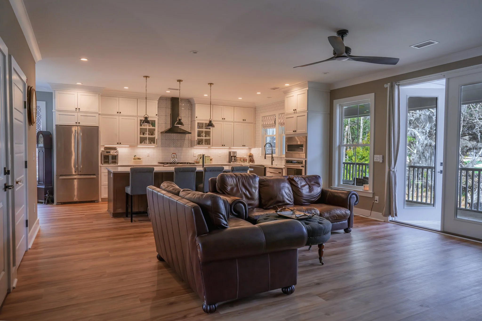 Open-concept living room and kitchen with hardwood floors, leather sofas, white cabinetry, large windows, and a balcony outside.