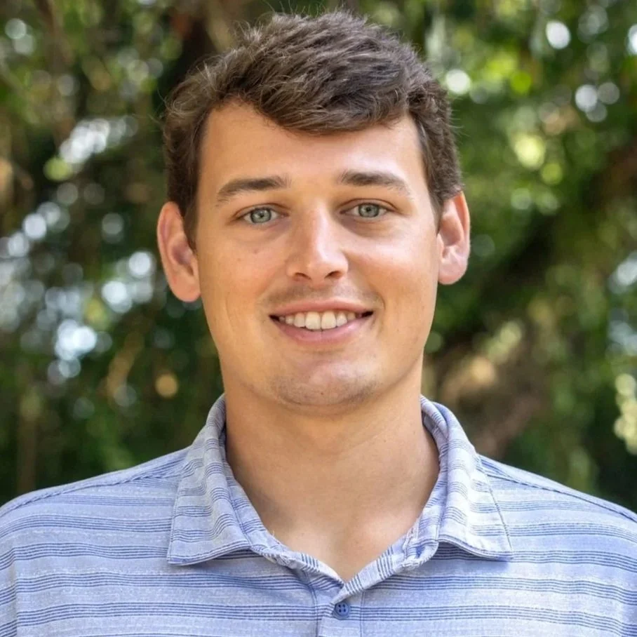 A young man with short brown hair, blue eyes, and light skin, smiling in front of a background of trees and greenery, wearing a blue striped collared shirt.