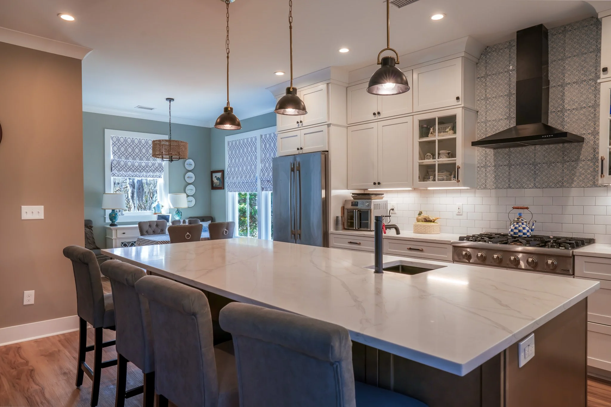 Modern kitchen with white cabinets, a large marble island, stainless steel appliances, black pendant lights, and a dining area with patterned window shades and decorative plates on the wall.