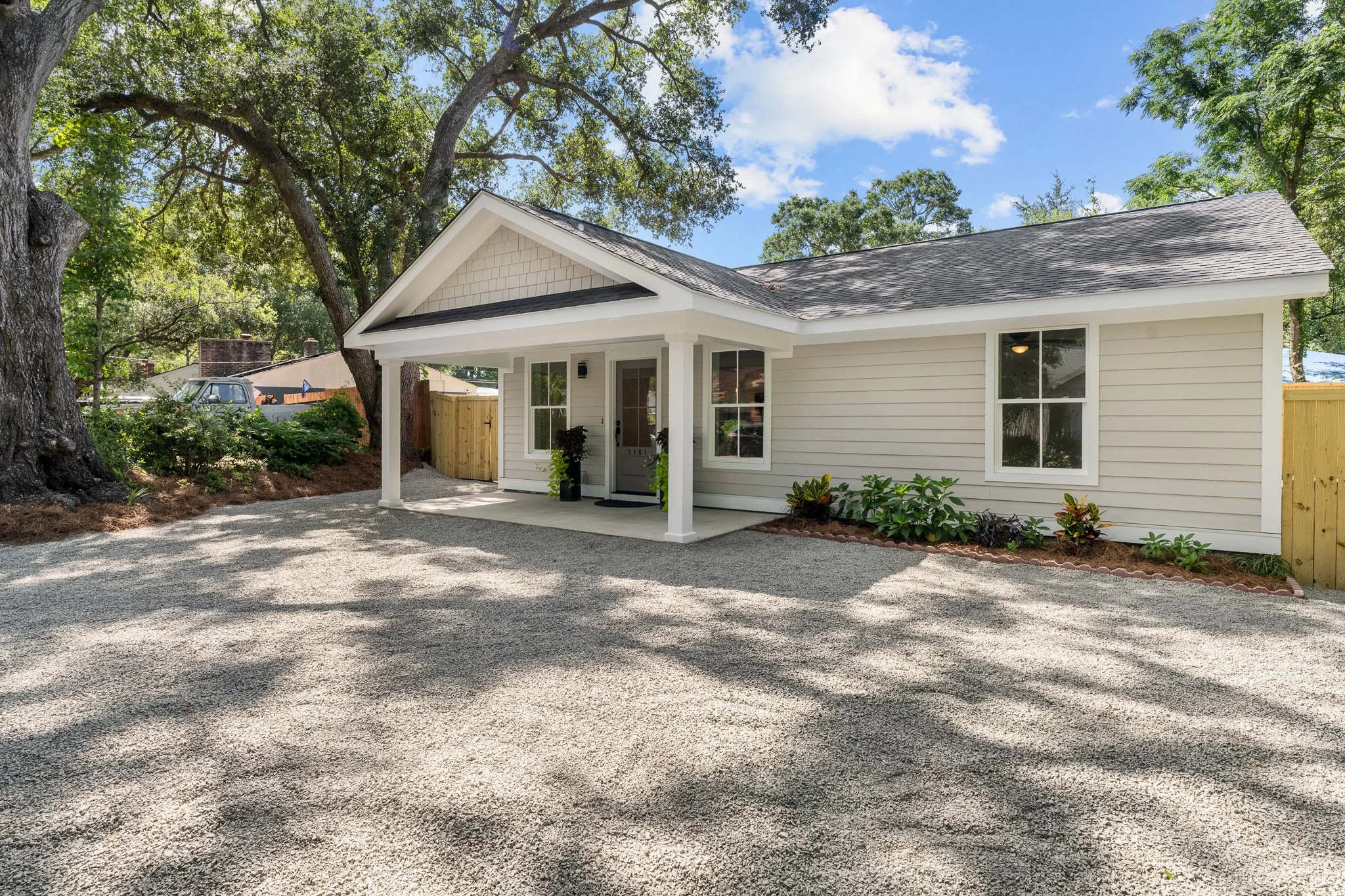 Front view of a house with a gravel driveway, beige siding, a small porch, and a garden bed with plants and shrubs, surrounded by trees and a wooden fence.