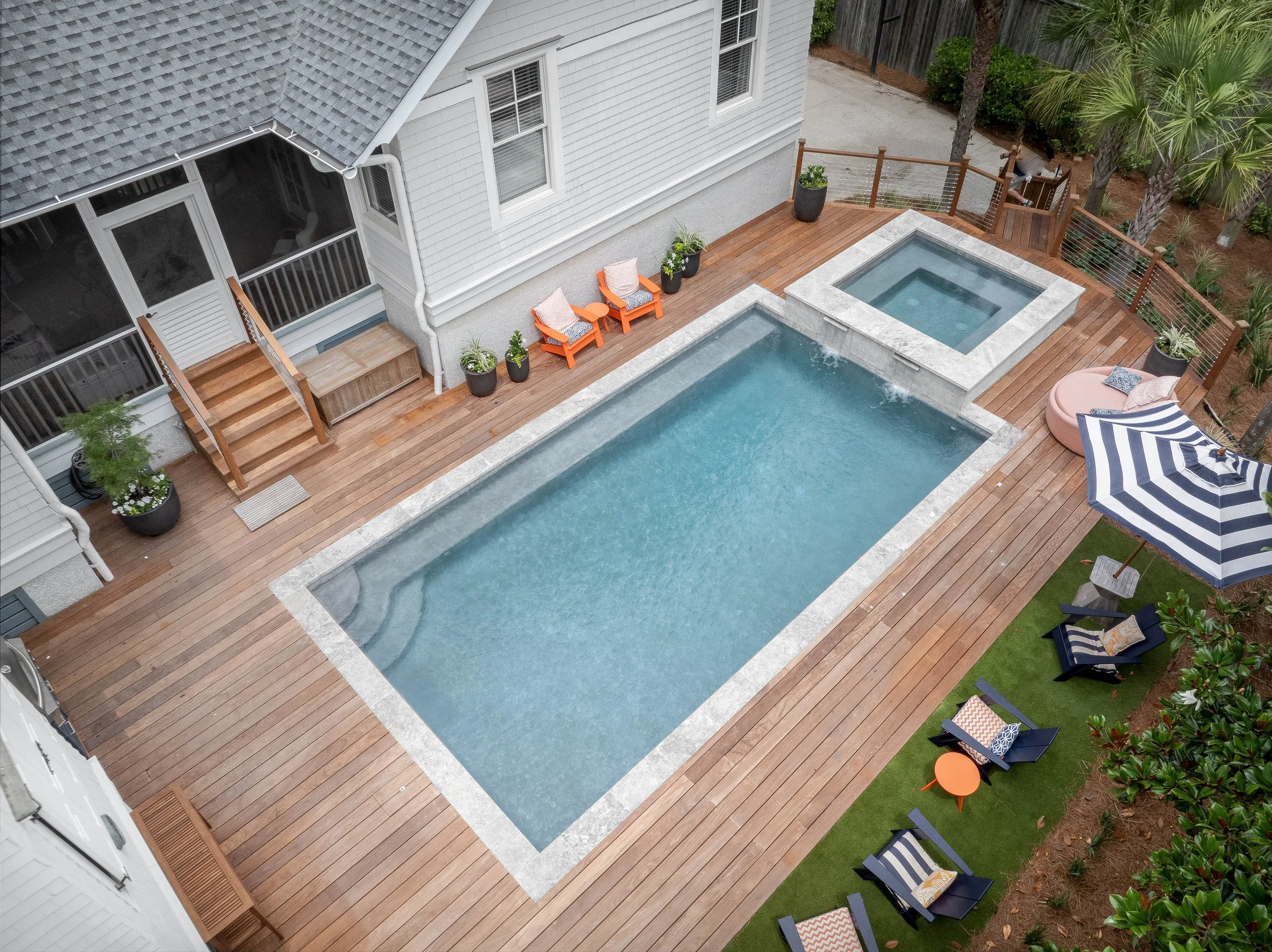 A backyard with a rectangular swimming pool, hot tub, wooden deck, and outdoor furniture including chairs, tables, and umbrellas.