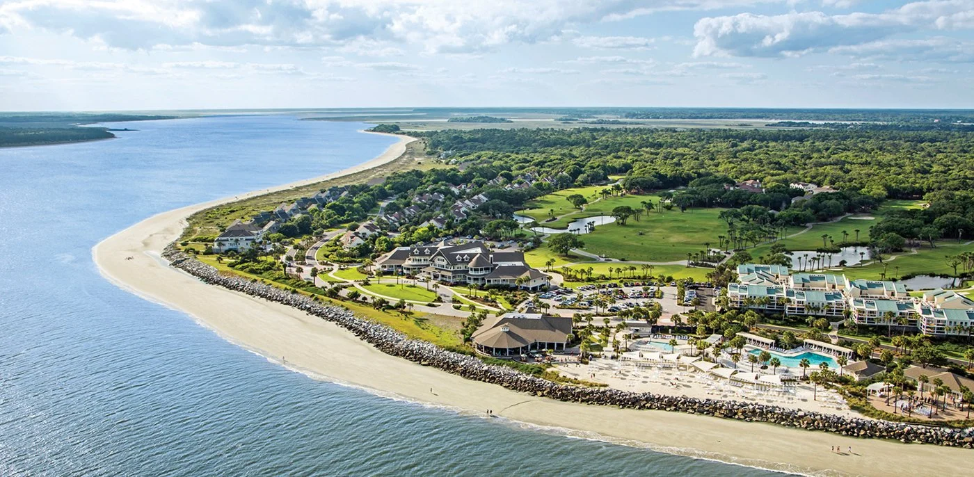 Aerial view of a resort along a beach with a marina, green lawns, a pool area, and residential buildings near a river or bay, with a forest in the background.