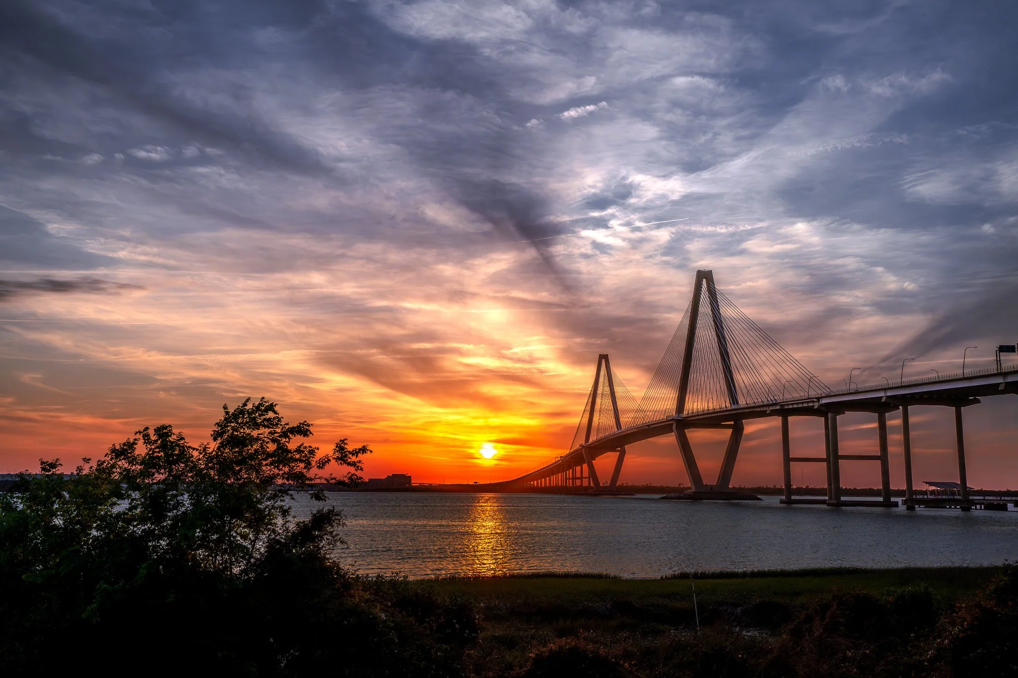 Sunset over a bridge spanning a river, with clouds in the sky and silhouettes of trees in the foreground.