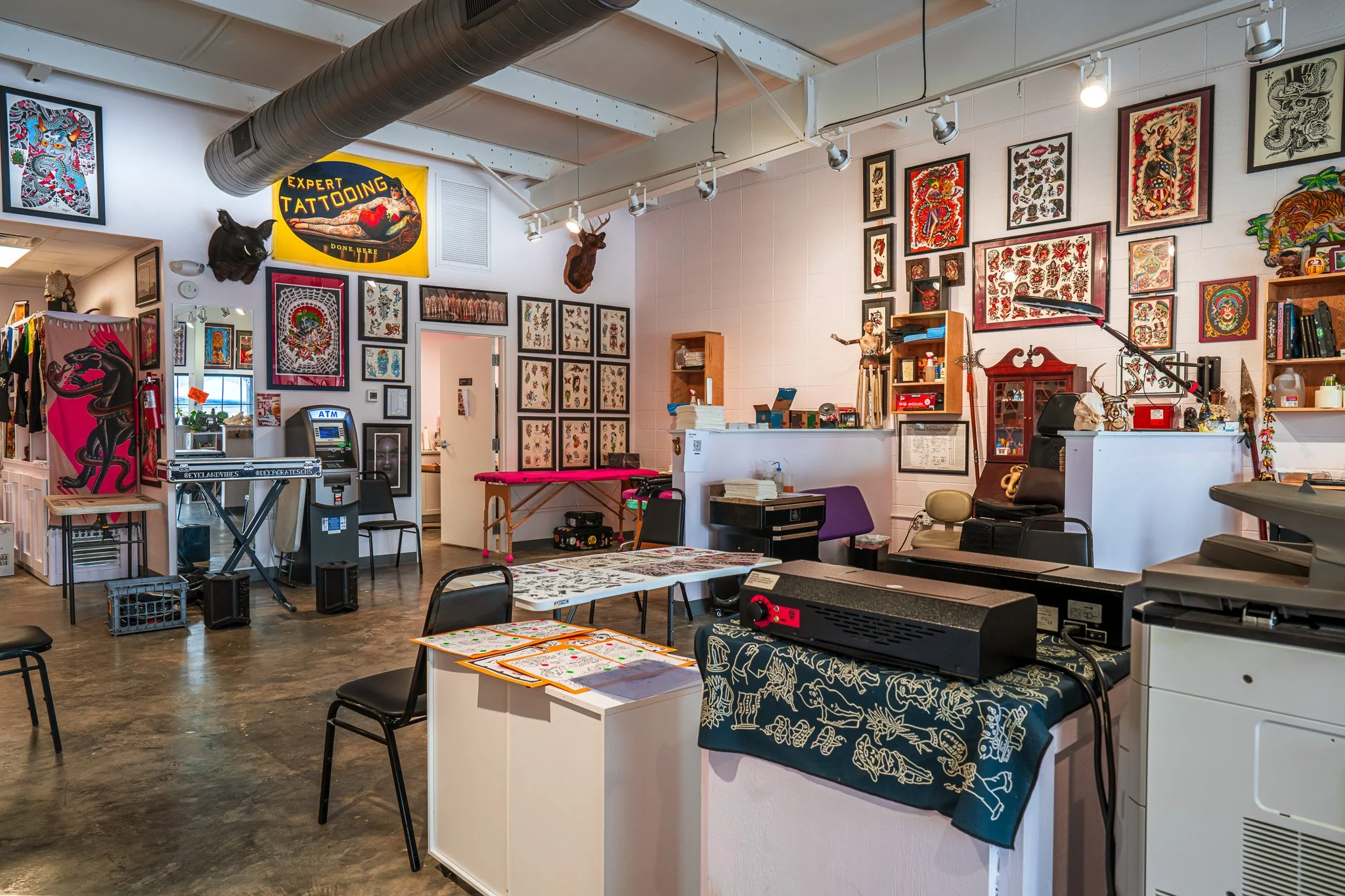 An interior view of a tattoo parlor with framed artwork, animal head mounts, and tattoo supplies on the walls, including a tattoo machine on a counter, a massage table, and chairs.