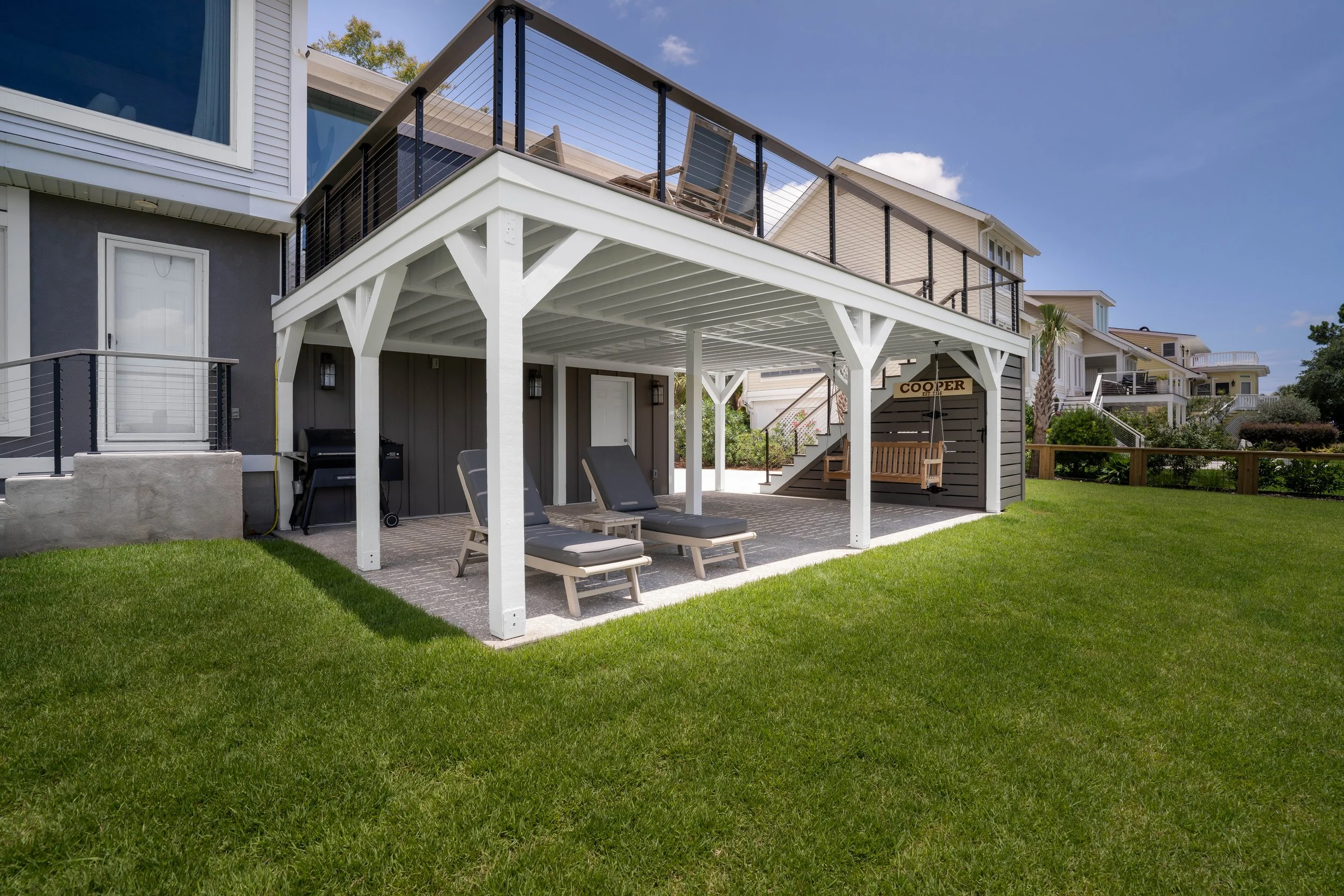 Backyard patio with under-deck area featuring lounge chairs, a grill, a wooden swing, and a sign that says 'Cooper' on a wall, with green grass and neighboring houses in the background.
