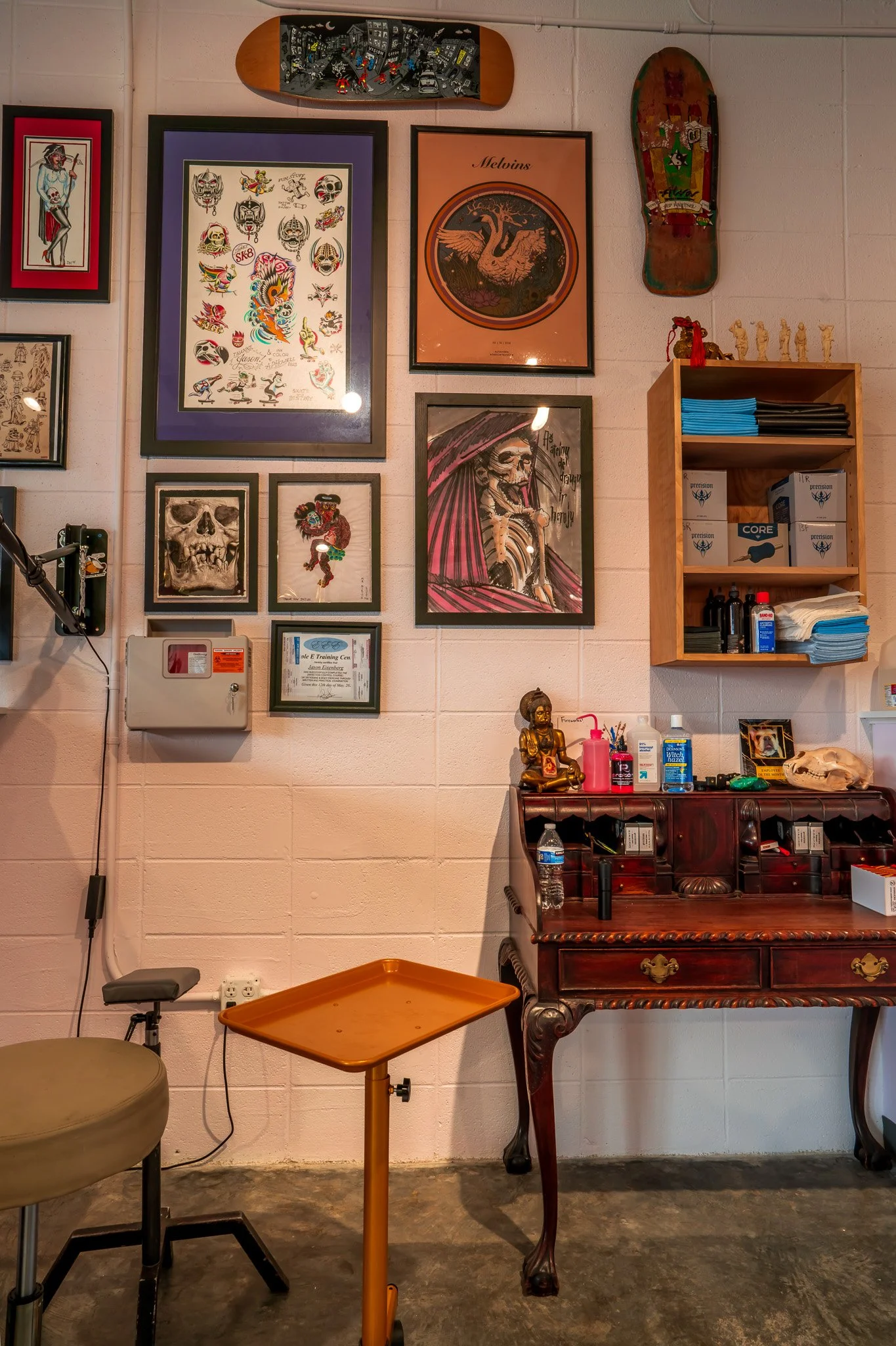 An art gallery wall with various framed posters and artwork, including skulls, a dragon, and a pirate. A skateboard with cityscape art is mounted above the frames. To the right is a wooden shelf with tissues, boxes, and miscellaneous items. Below the