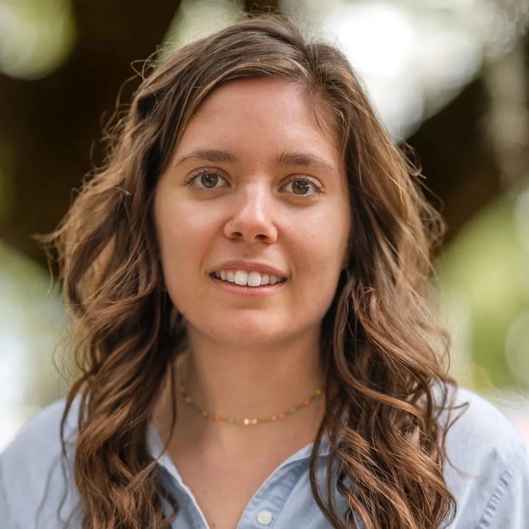Portrait of a young woman with wavy brown hair, brown eyes, and a bright smile, wearing a light blue shirt and a colorful beaded necklace outdoors with blurred green foliage in the background.