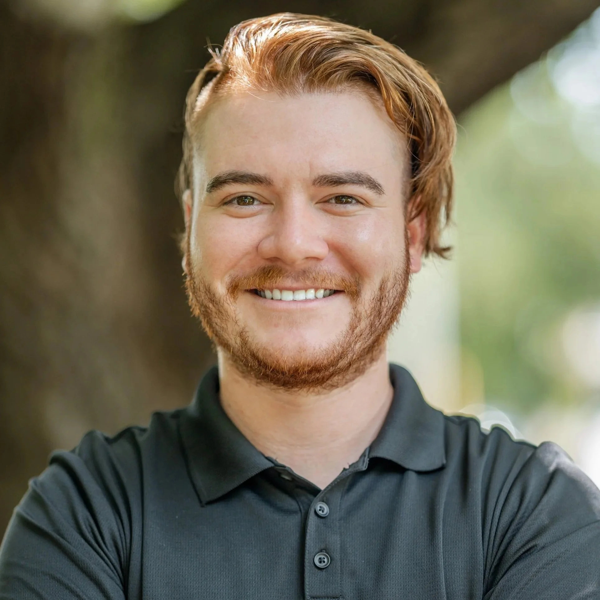 Young man with reddish-brown hair and a beard smiling outdoors in front of a blurred background.