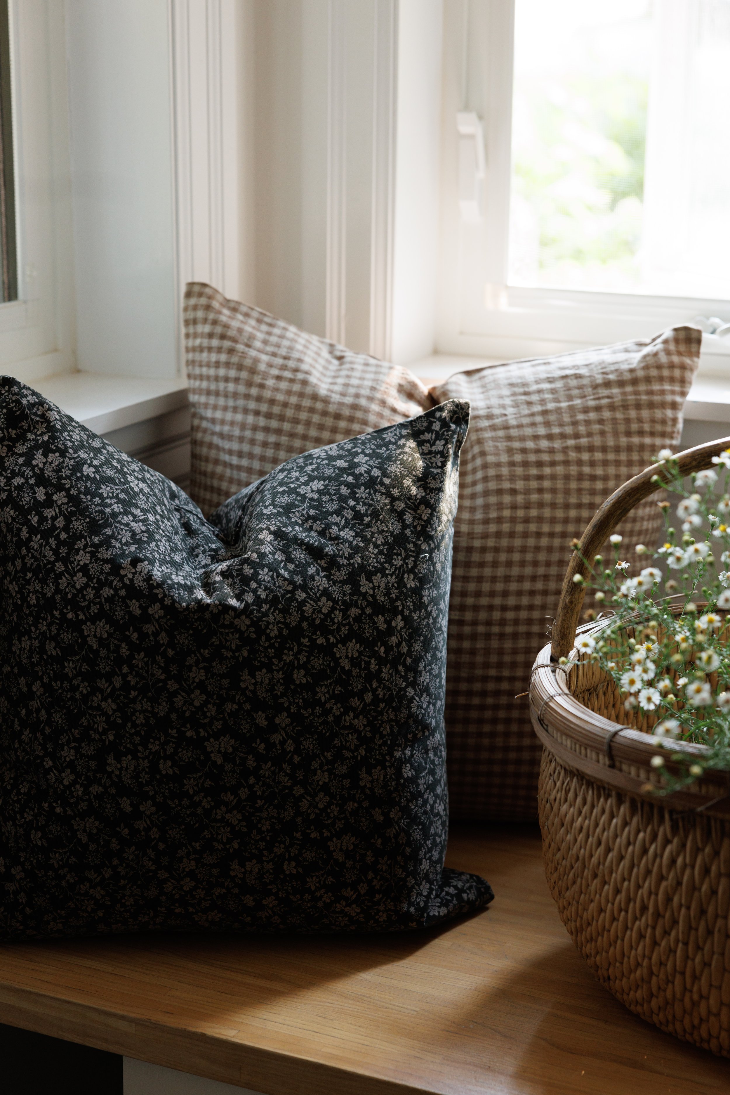 Sunlit window sill with gingham and floral pillows and a wicker basket of white flowers