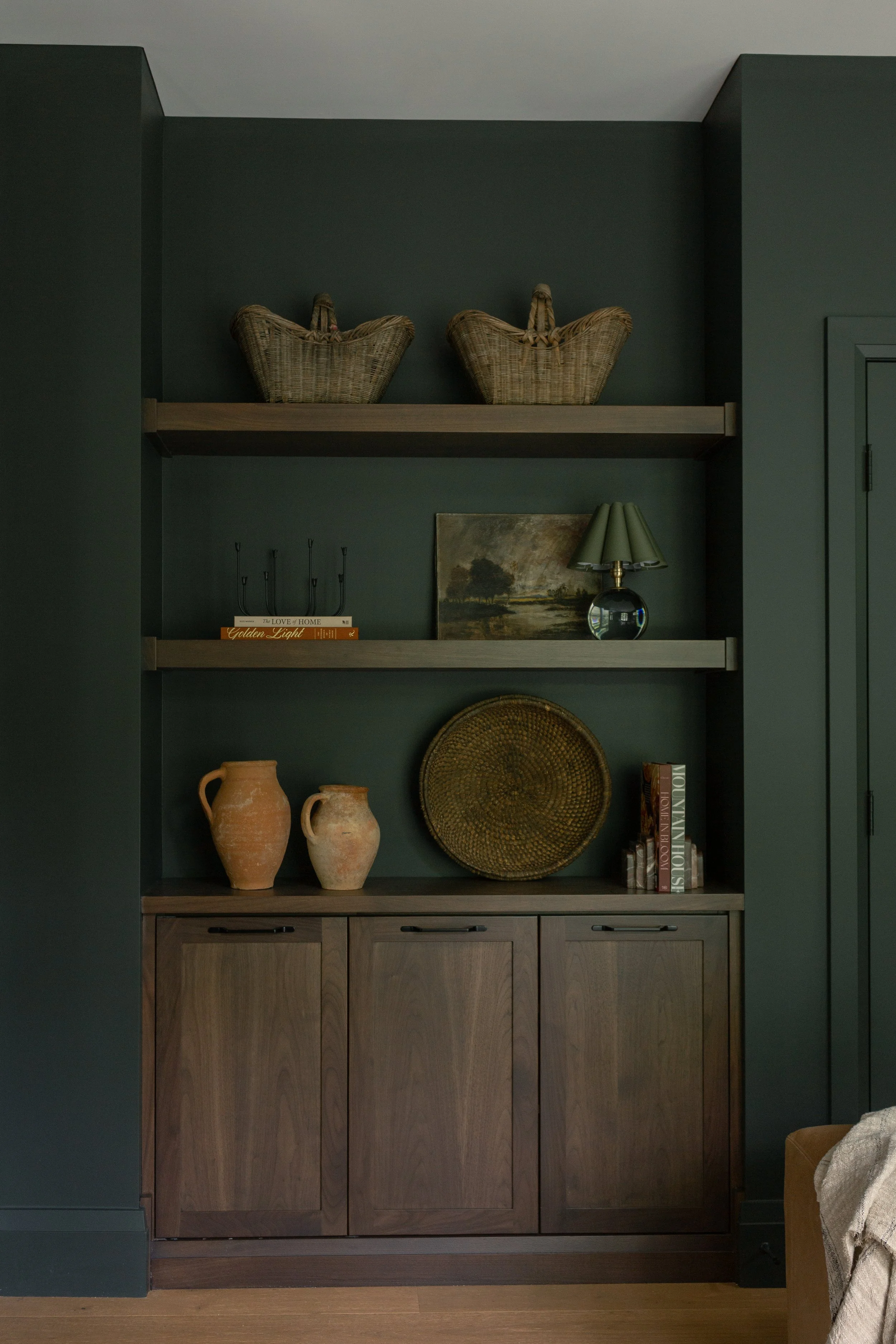 A wooden cabinet with decorative pottery, a woven basket, and a stack of books on top, against a dark green wall with shelves holding wicker baskets, books, a landscape painting, and a small glass lamp with a green shade. Interior Design Guelph