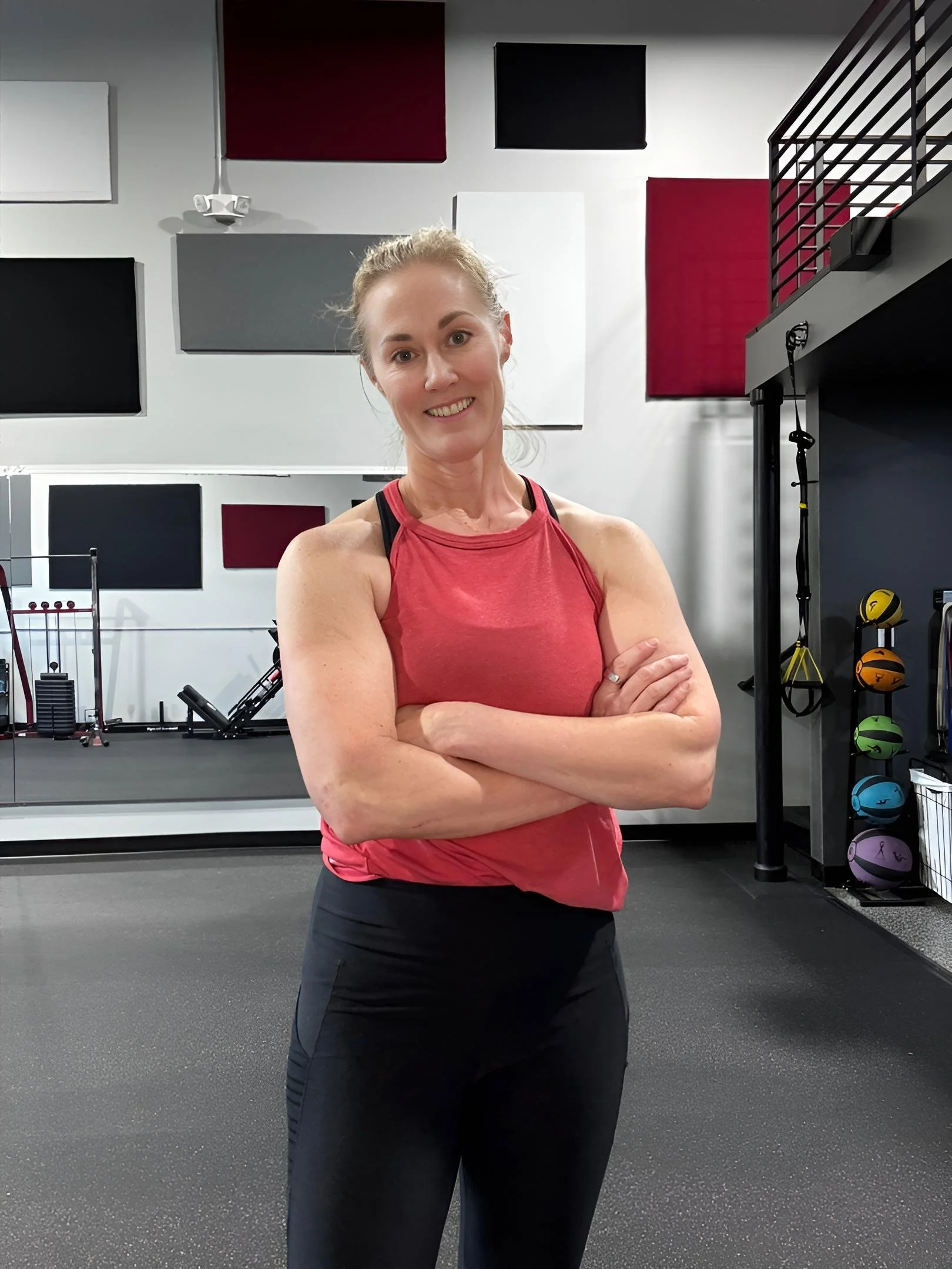 A woman in workout attire standing inside a gym with arms crossed, smiling at the camera, with exercise equipment and soundproofing panels on the wall behind her.
