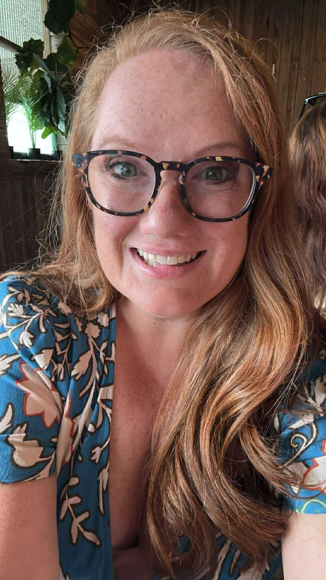 A woman with long, wavy red hair and glasses with a tortoise shell pattern, smiling indoors with plants and wooden walls in the background.