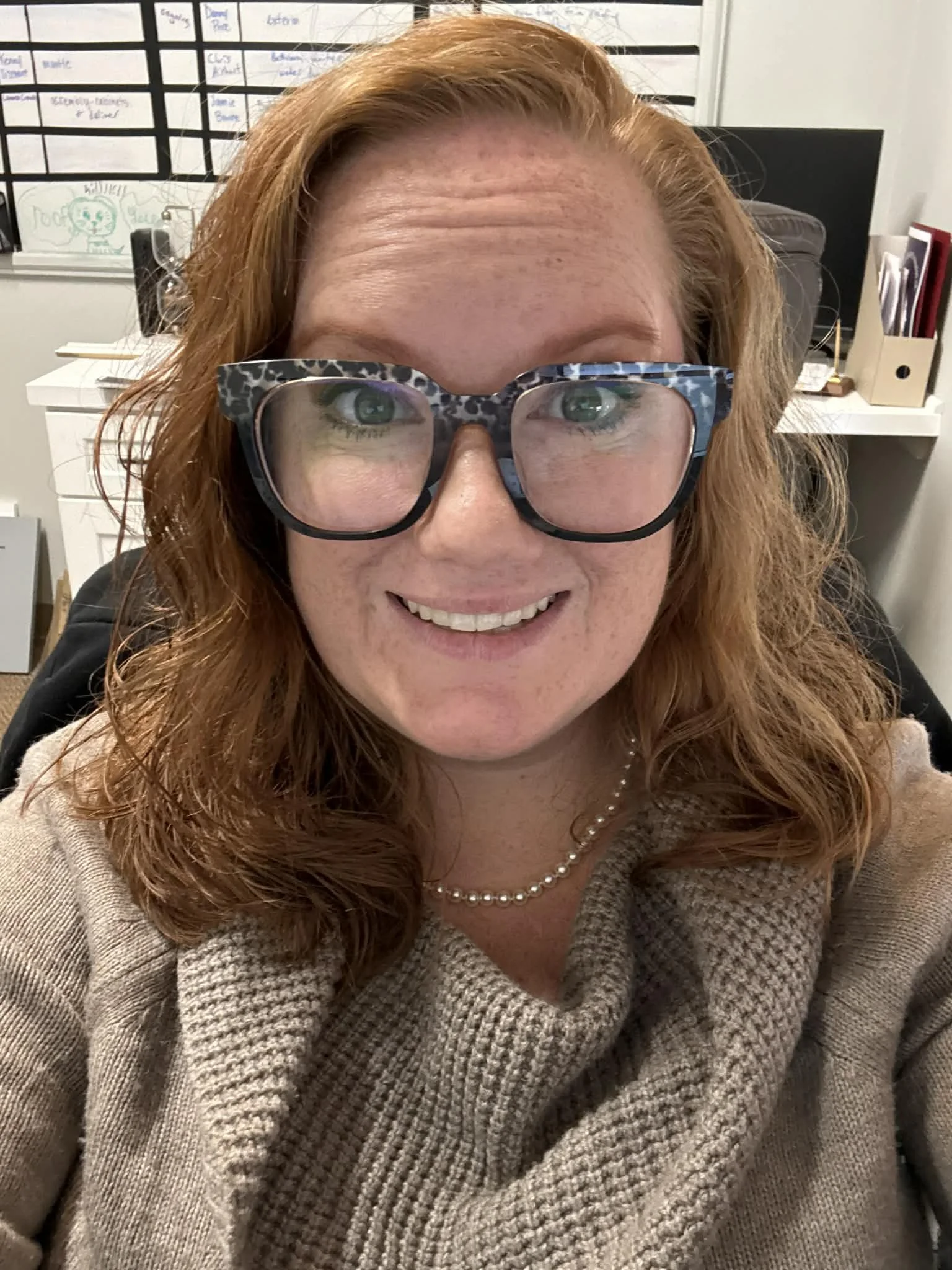 A woman with red hair, wearing glasses with a leopard print frame, smiling and looking at the camera in an office setting.