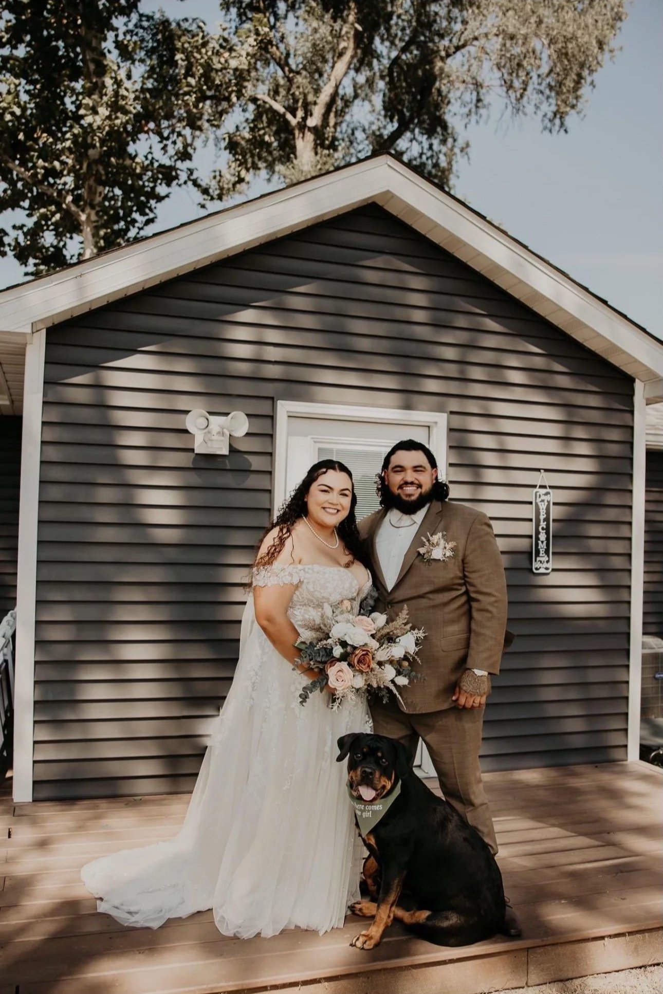 A newlywed couple standing in front of a dark gray house, smiling and posing with their dog. The bride is in a white wedding dress holding a bouquet, and the groom is in a brown suit with a boutonniere. Shadows of leaves fall on the house behind them.
