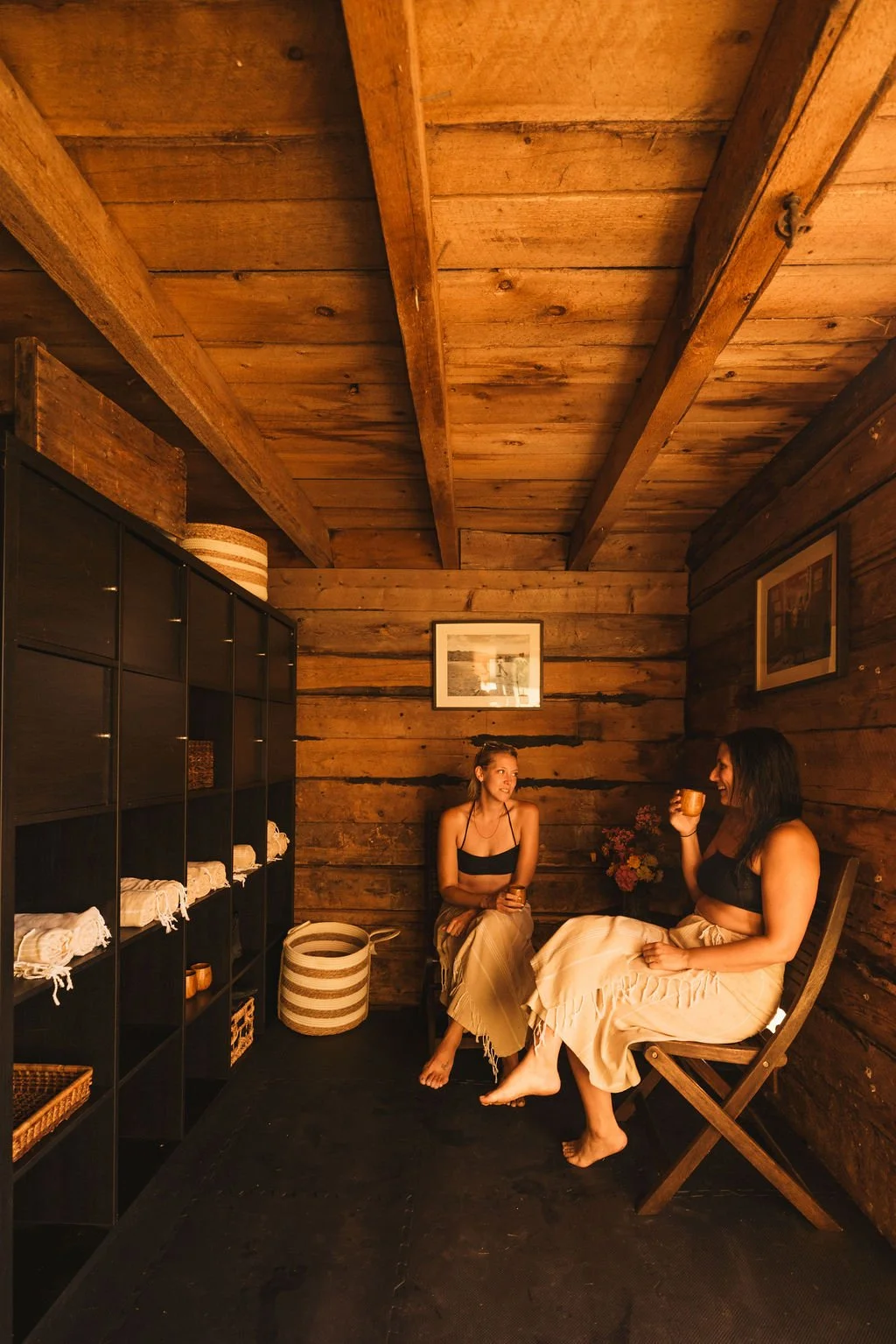 Two women sitting in a cozy wooden room, one is holding a mug, engaged in conversation, surrounded by shelves with towels and baskets, warm lighting.