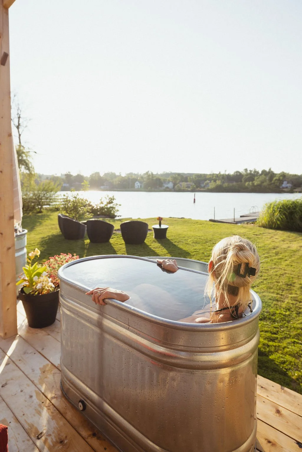 A woman with blonde hair in a hair clip relaxing in an outdoor ice tub by a lake, with patio furniture and flowering plants nearby, during sunset.