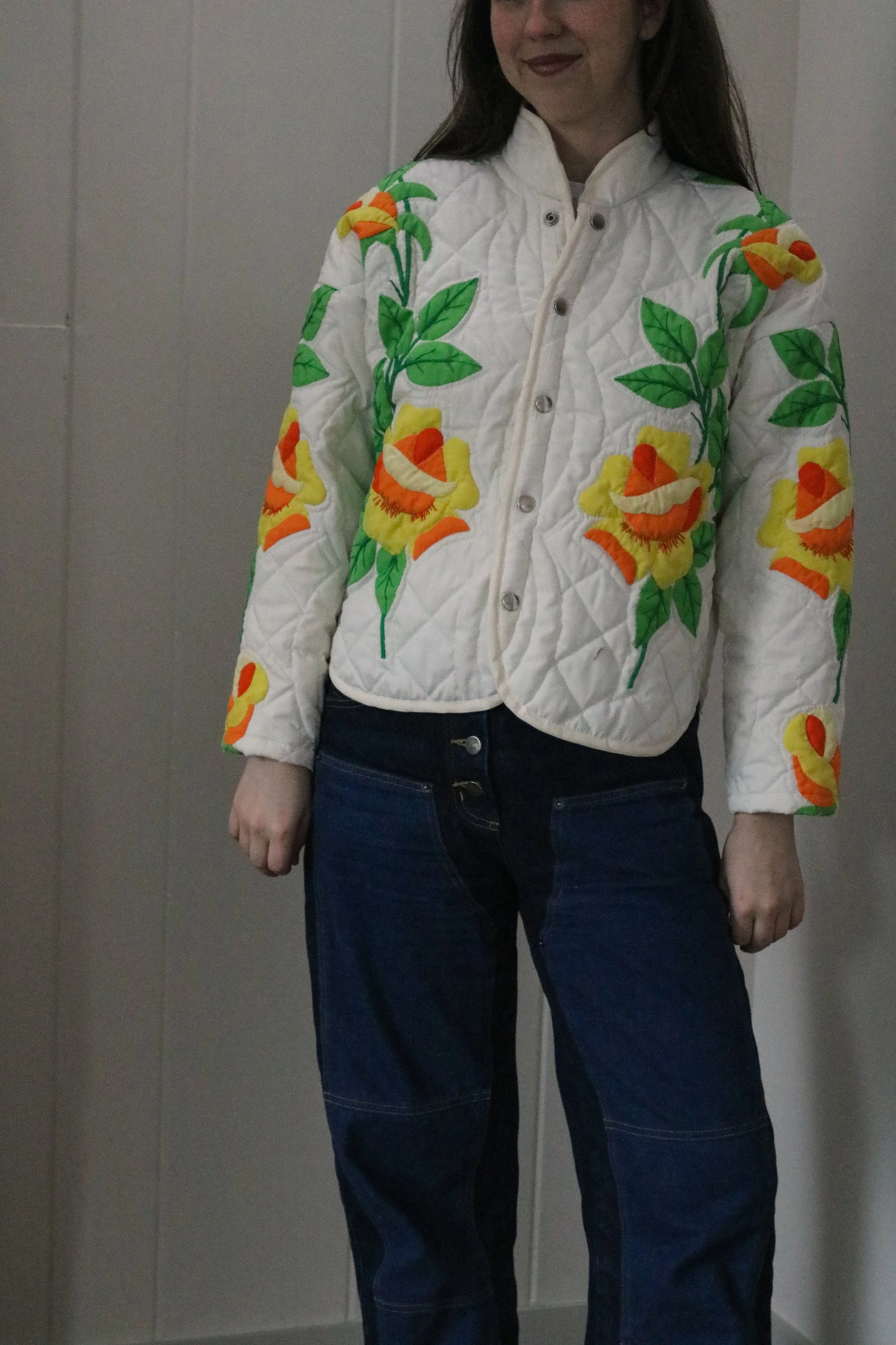 Person wearing a quilted white jacket with a floral pattern of large yellow and orange flowers and green leaves, standing against a plain wall.
