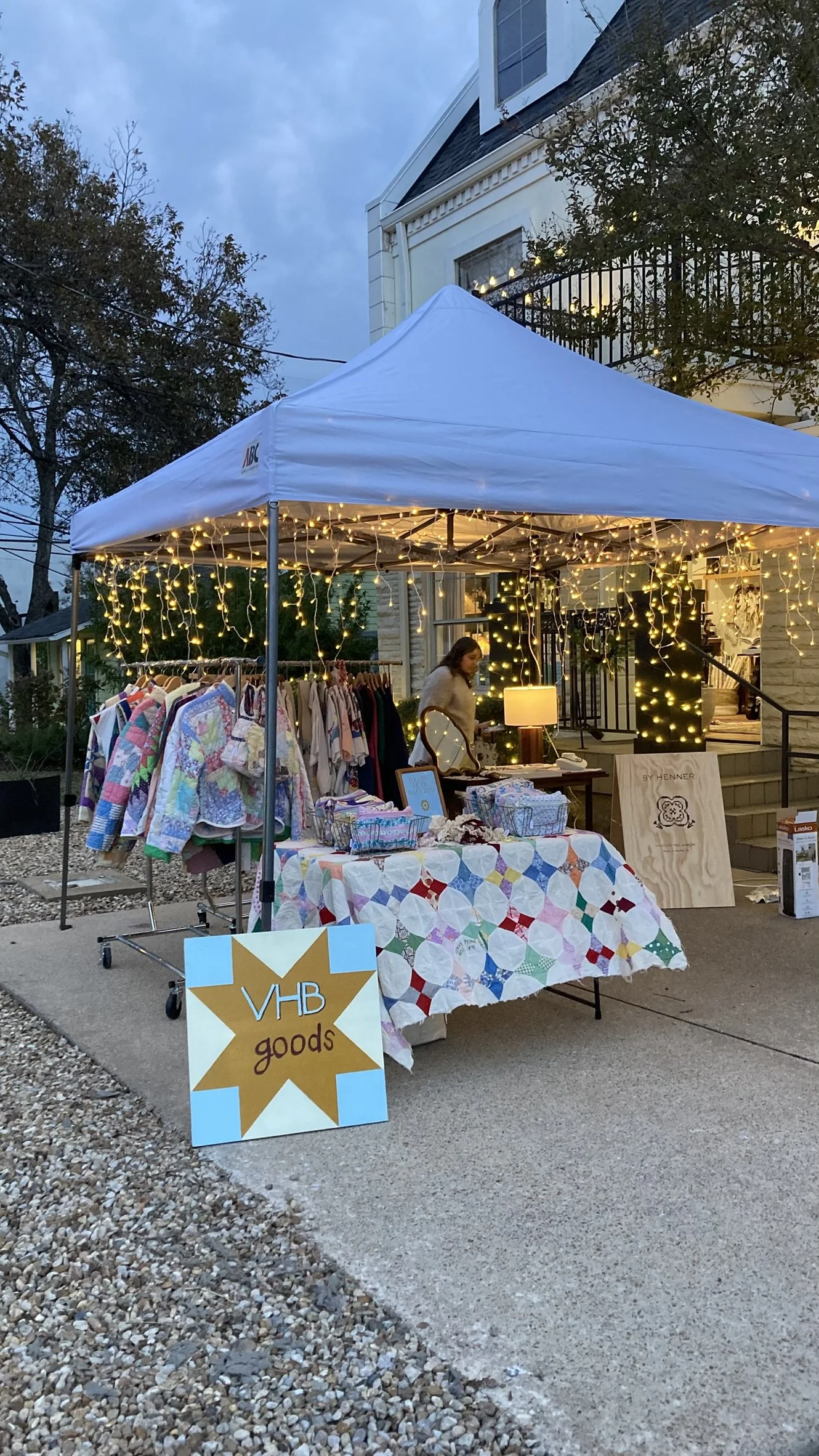 Outdoor yard sale booth under a white canopy with string lights, displaying colorful clothing, accessories, and decorative items on tables and racks, with a sign that reads 'VHB goods'.