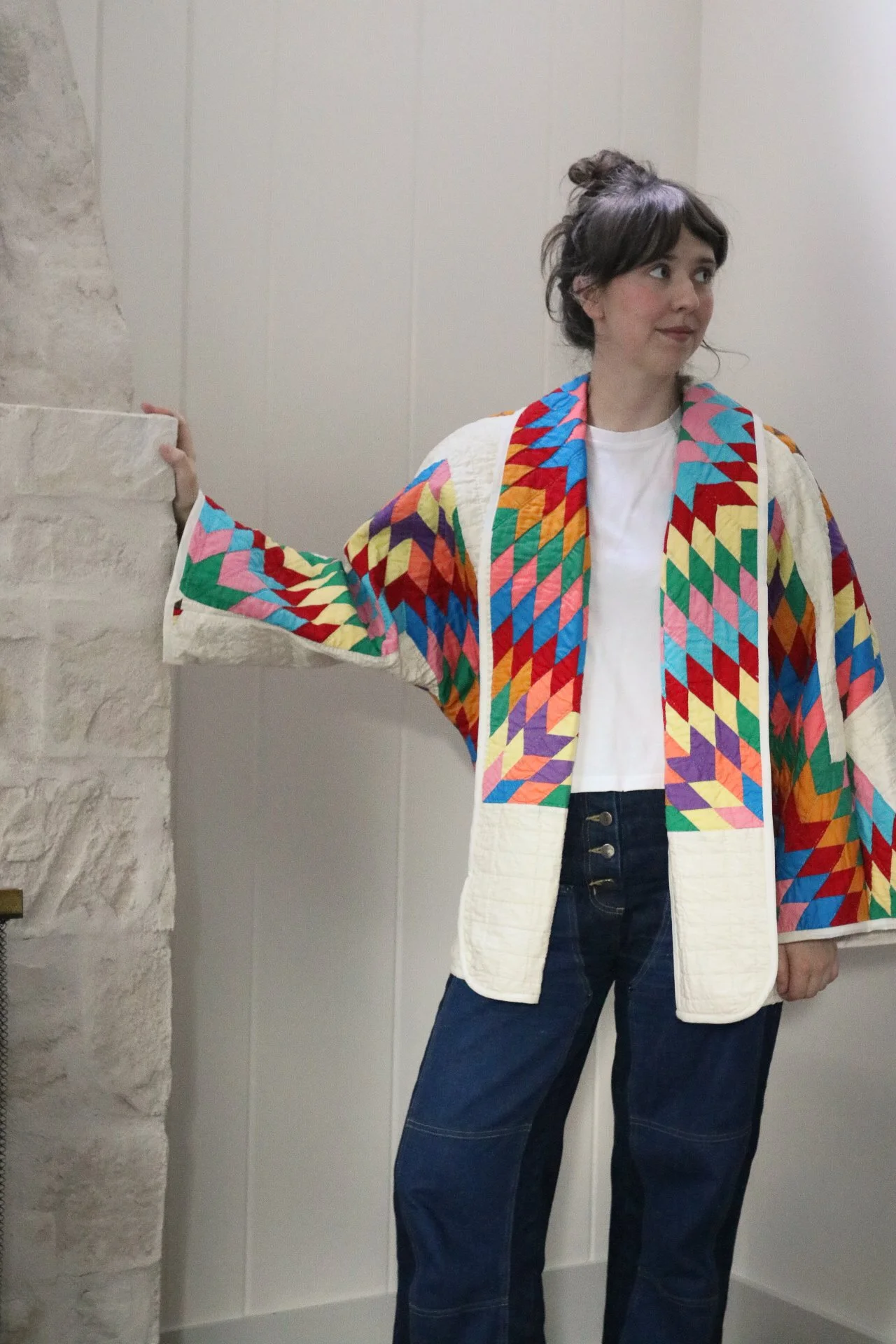 Woman with short, curly brown hair, wearing a colorful quilted jacket with geometric patterns, white T-shirt, and dark jeans, standing indoors against a light-colored wall with a stone fireplace on the left.