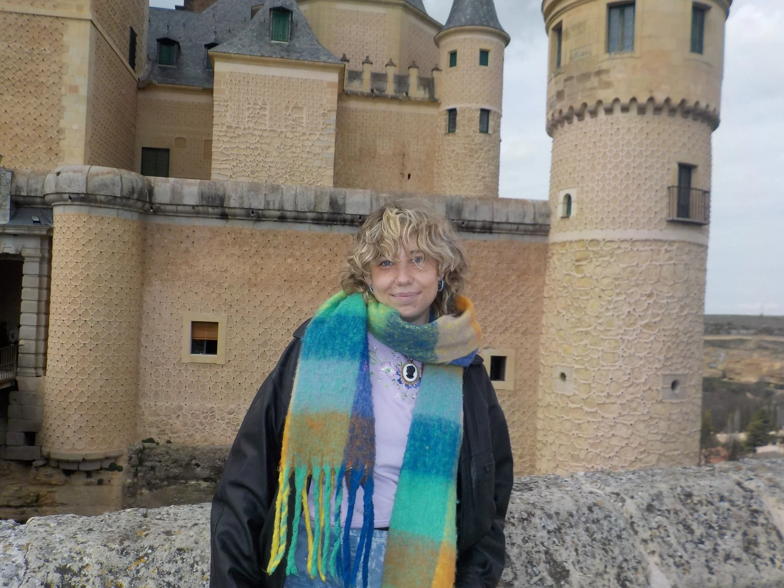 Person wearing a colorful scarf with short blonde hair, posing infront of a castle.