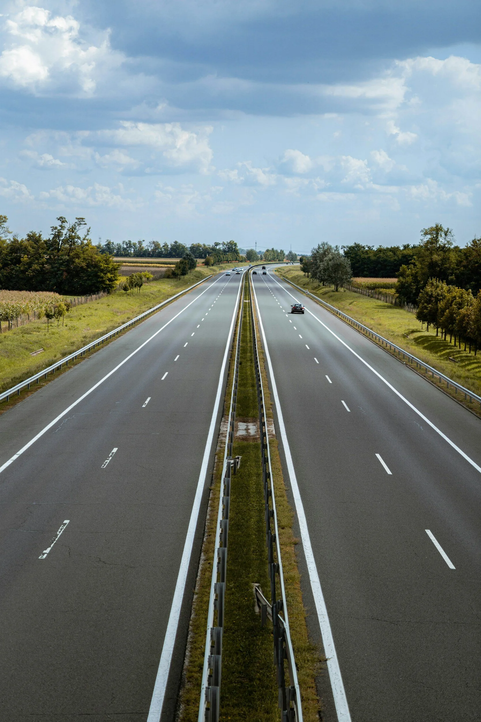 Empty highway with multiple lanes, a grassy median, and trees along the sides, under a partly cloudy sky.