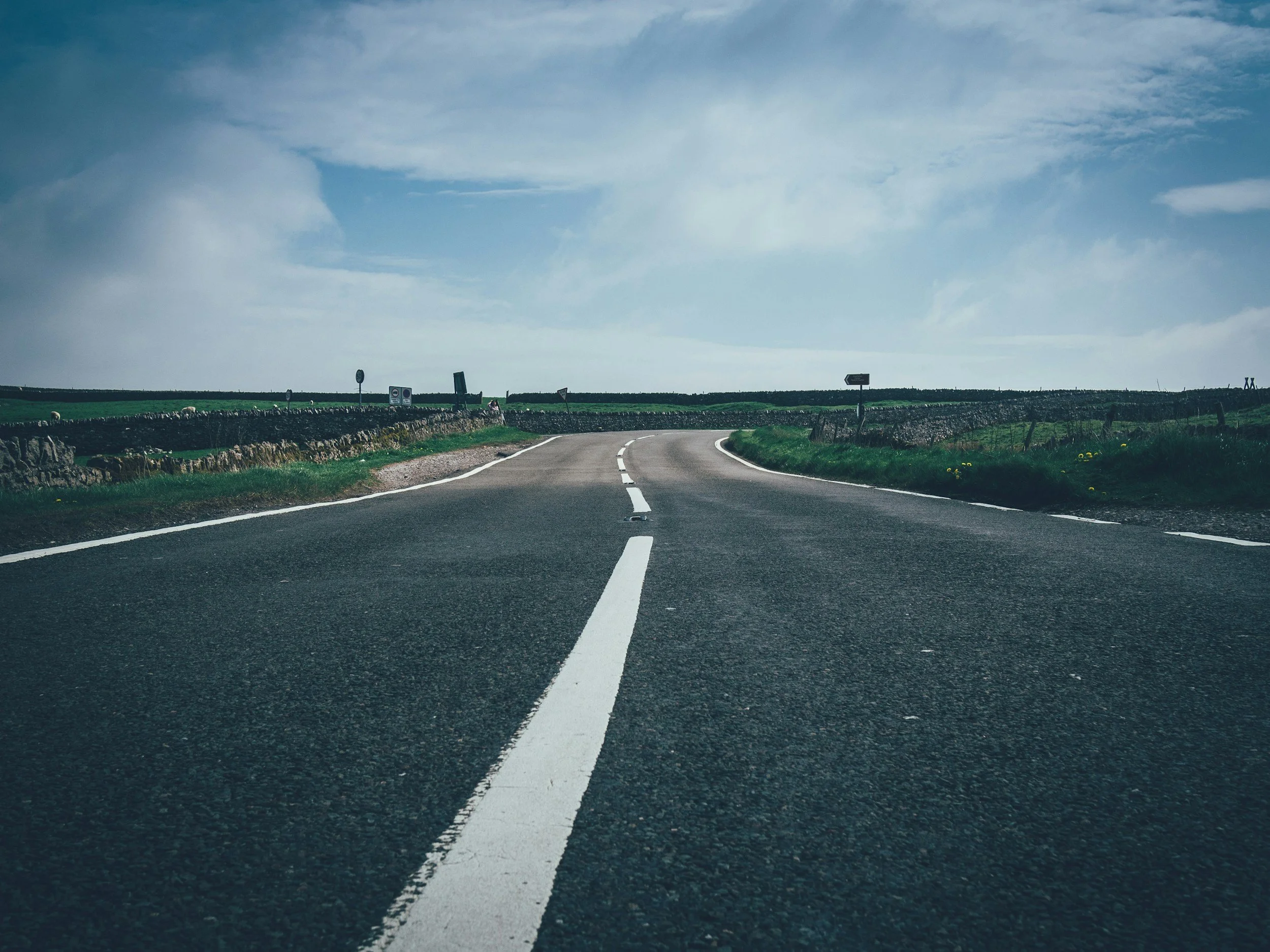 A rural two-lane asphalt road with white dashed lines, curving to the right, in a countryside landscape with green fields, stone walls, and cloudy sky.