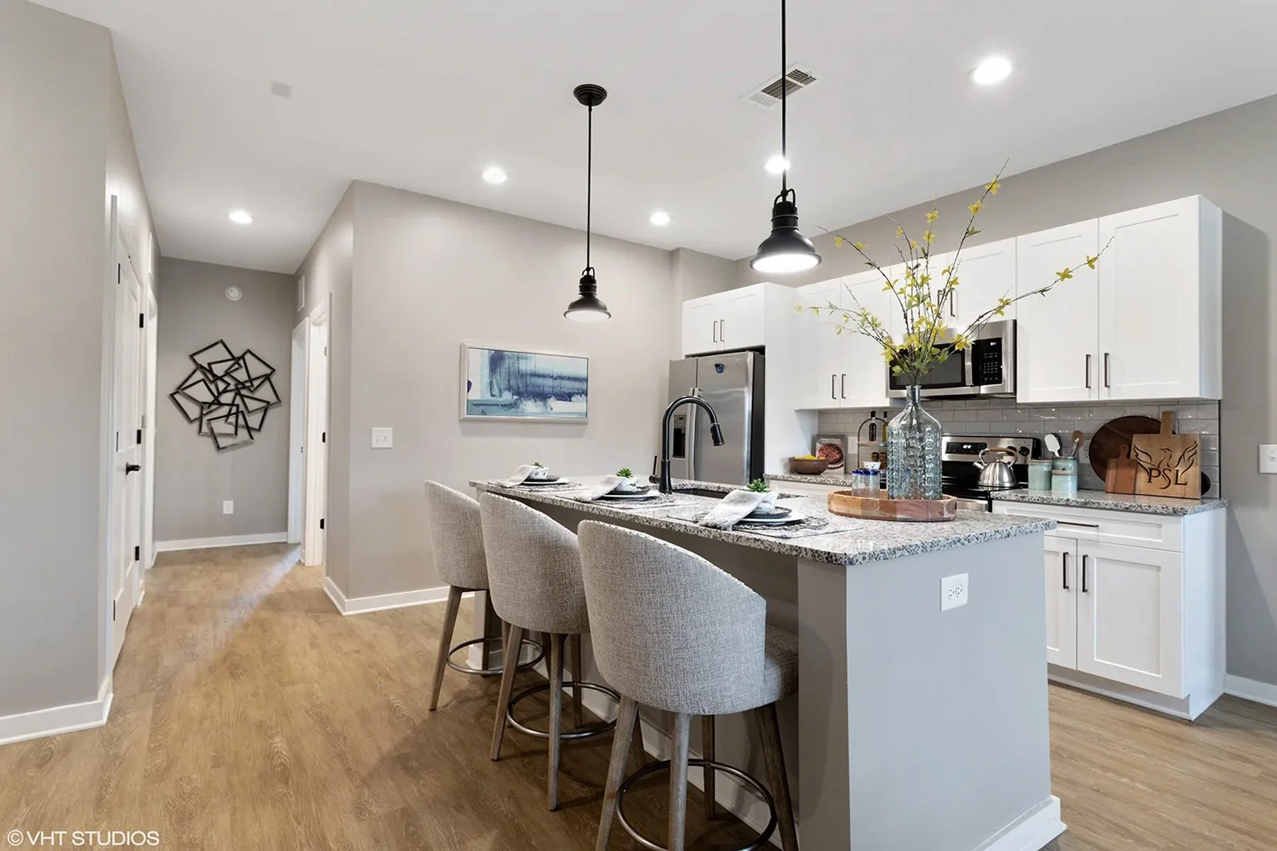 Modern kitchen with white cabinets, granite countertops, stainless steel appliances, and a breakfast bar with three beige upholstered chairs. There is a large glass vase with yellow flowering branches on the counter, decorative items, and a framed artwork on the light gray wall. The ceiling has recessed lighting and two black pendant lights hanging over the island.
