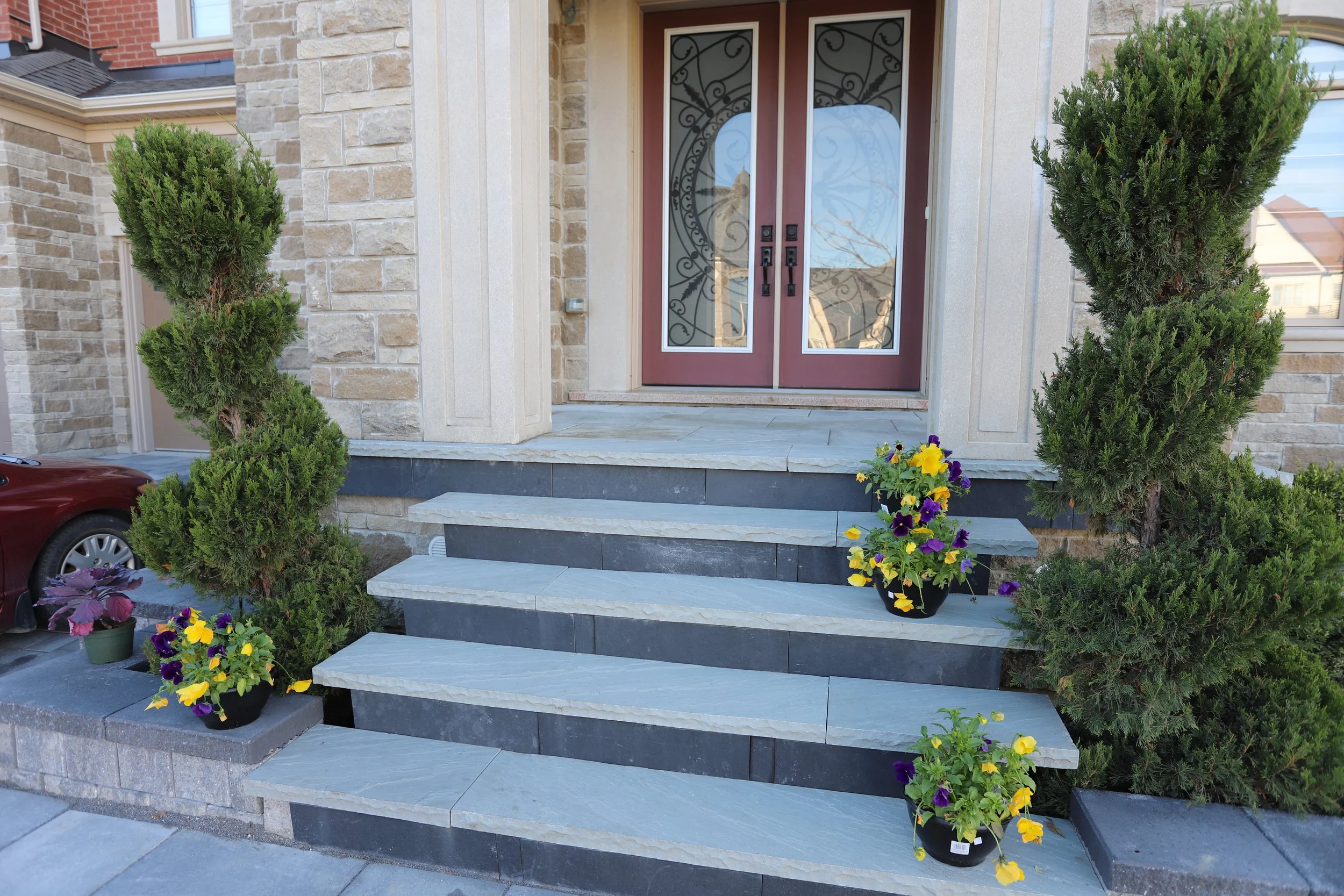 Stone steps leading to an ornate front door, flanked by potted plants with green shrubs and pansies in purple and yellow, set against a brick exterior wall.