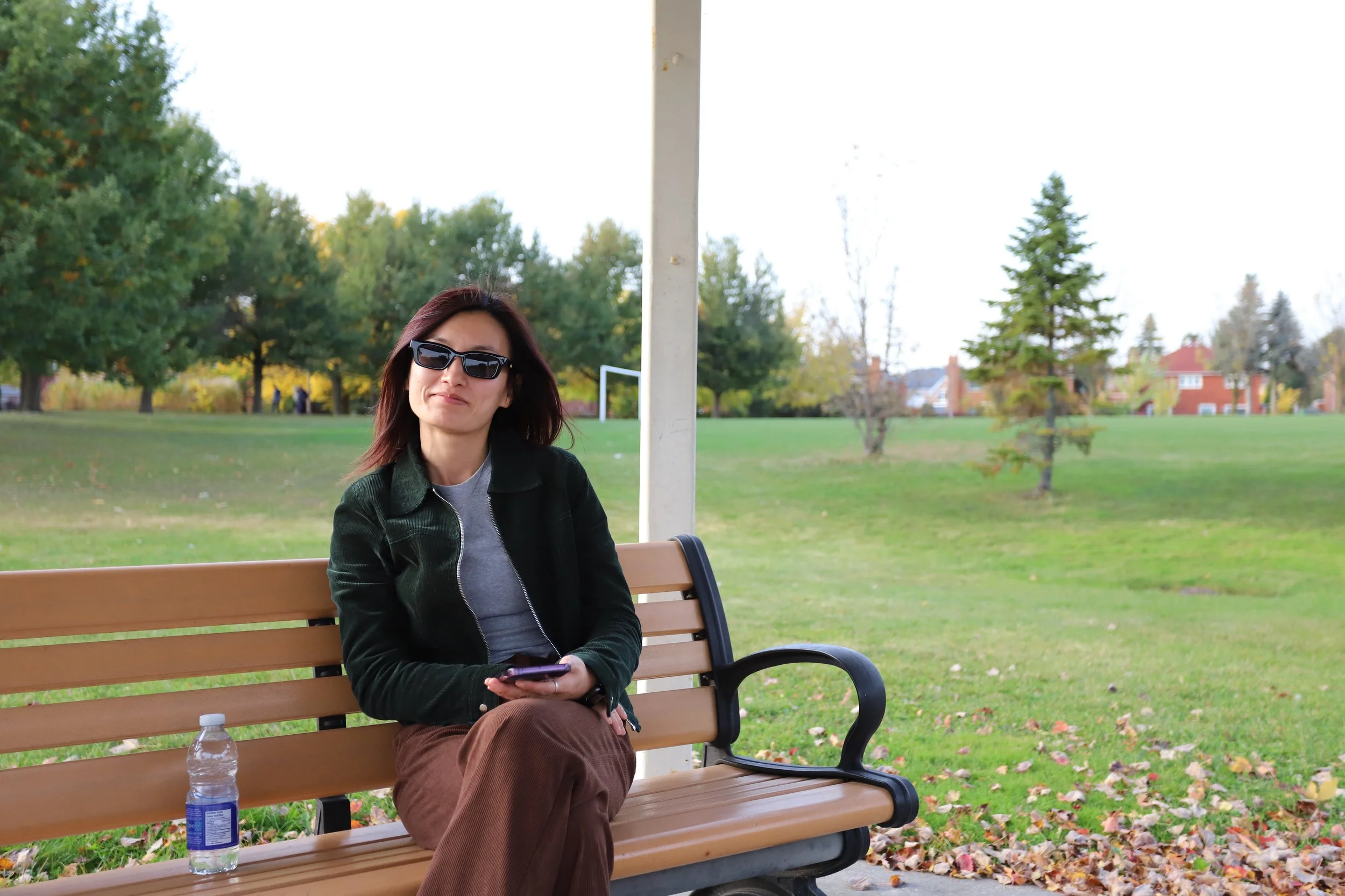 A woman wearing sunglasses sits on a park bench with a water bottle beside her. She is holding a phone and looking at the camera. The background has trees and a grassy field.